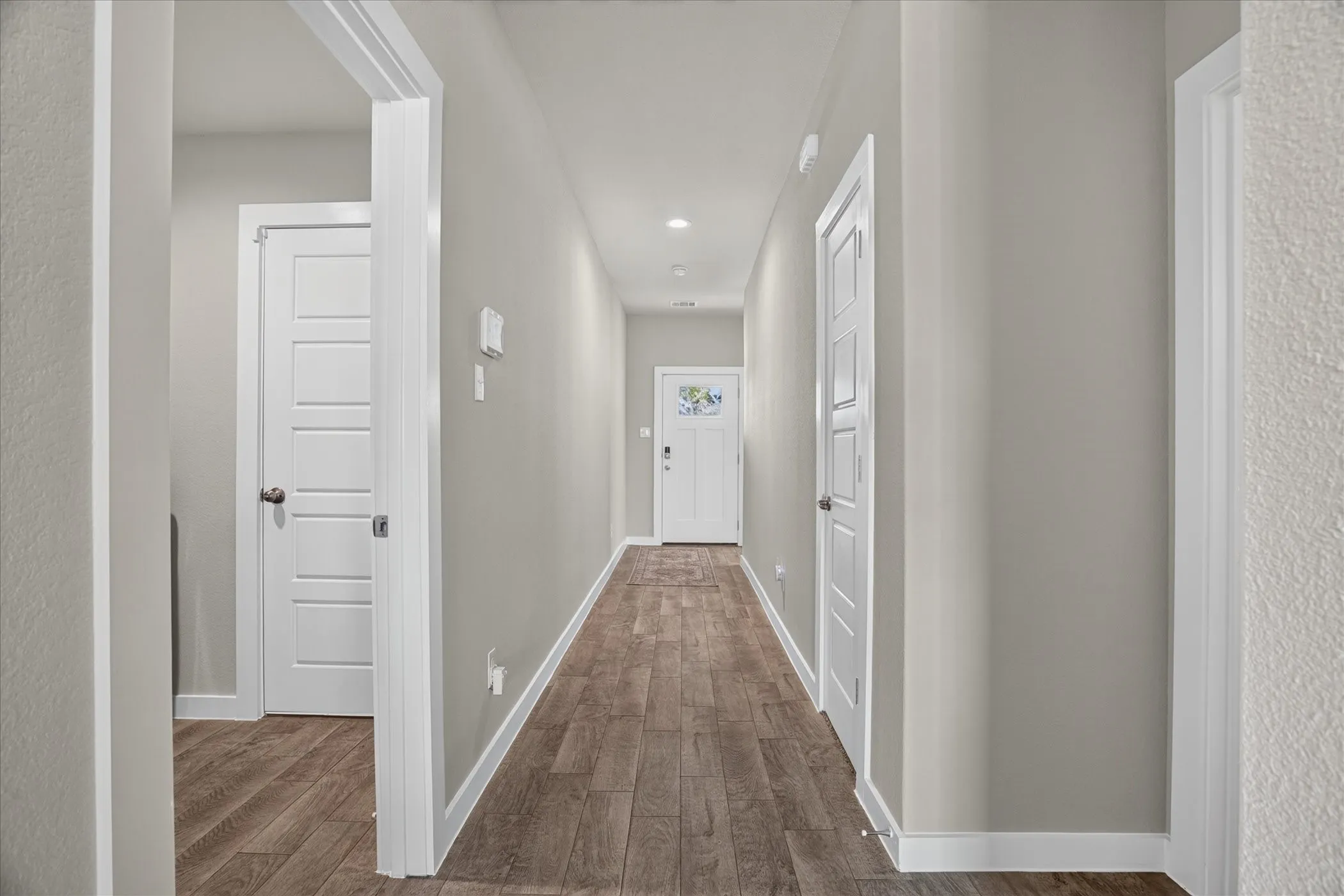 Hallway with wood-finish flooring, light gray walls, white trim, and recessed lighting, to the far right off front door is 2 bedrooms and spare bathroom, directly to the right is another bedroom and to the left is the utility room.