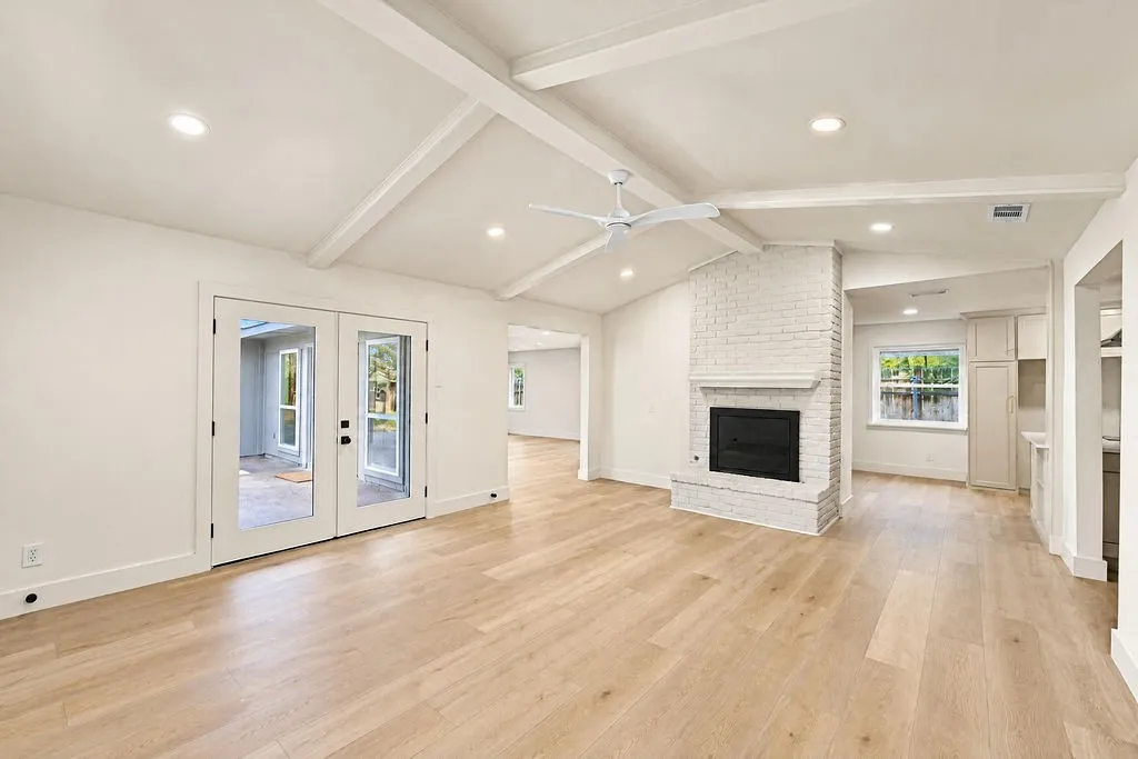 Vaulted ceiling with exposed beams, wood-finish flooring, white brick fireplace with hearth, recessed lighting, and white French doors