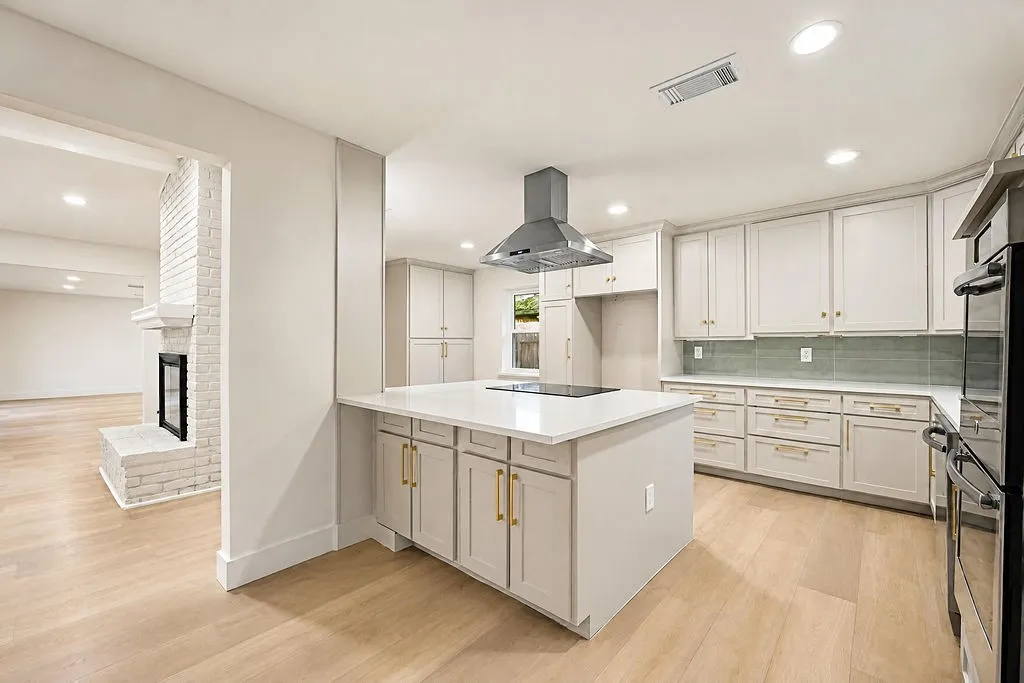 Modern kitchen with a large island, stainless steel range hood, light wood-finish flooring, and light-toned cabinetry with gold-tone hardware