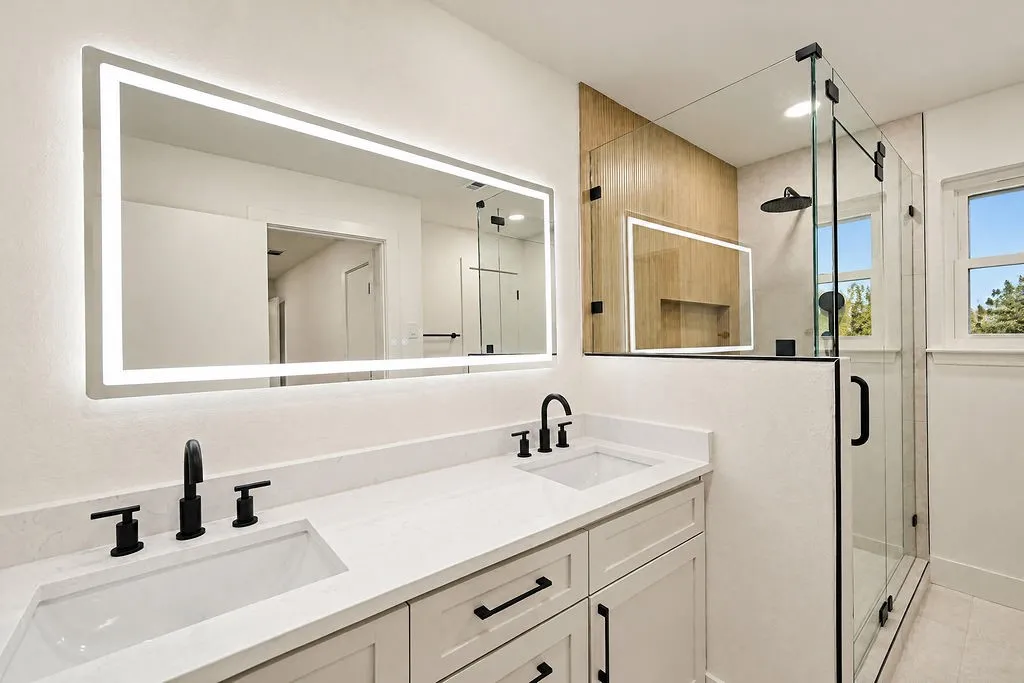 Dual vanity with integrated sinks and matte black fixtures, backlit mirror, glass-enclosed shower with wood-finish accent wall, and a rain shower head