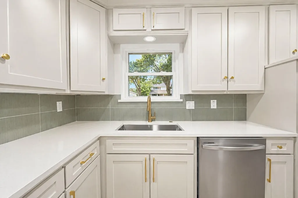 White shaker-style cabinetry with gold-tone hardware, light-toned countertops, and a stainless steel dishwasher