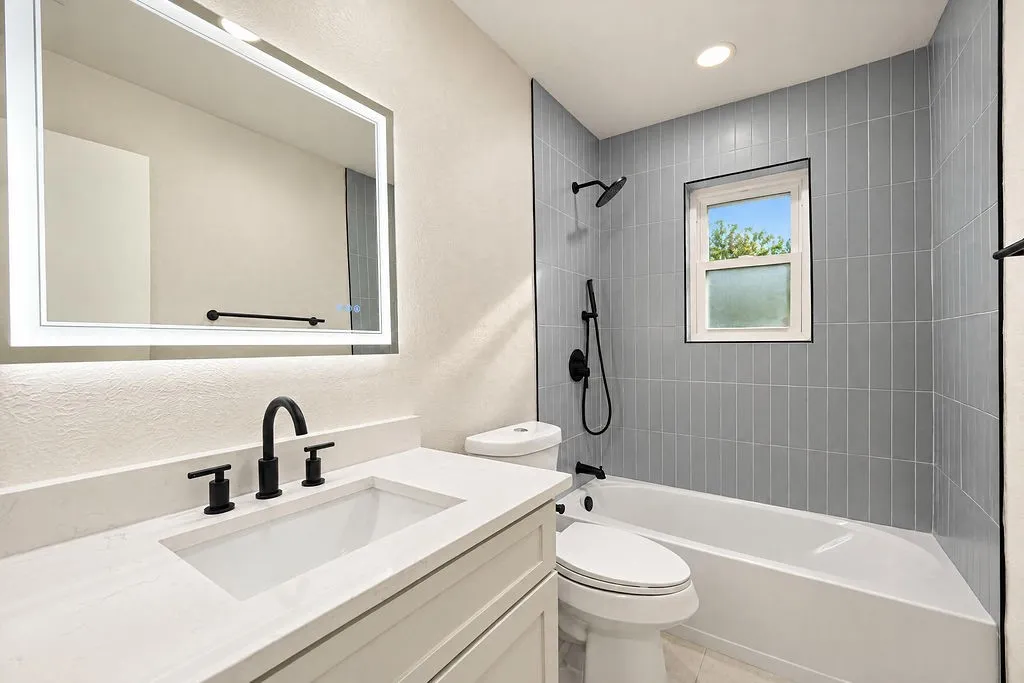 Bathroom featuring a vanity with an integrated sink, backlit mirror, matte black fixtures, and a tiled shower with a window