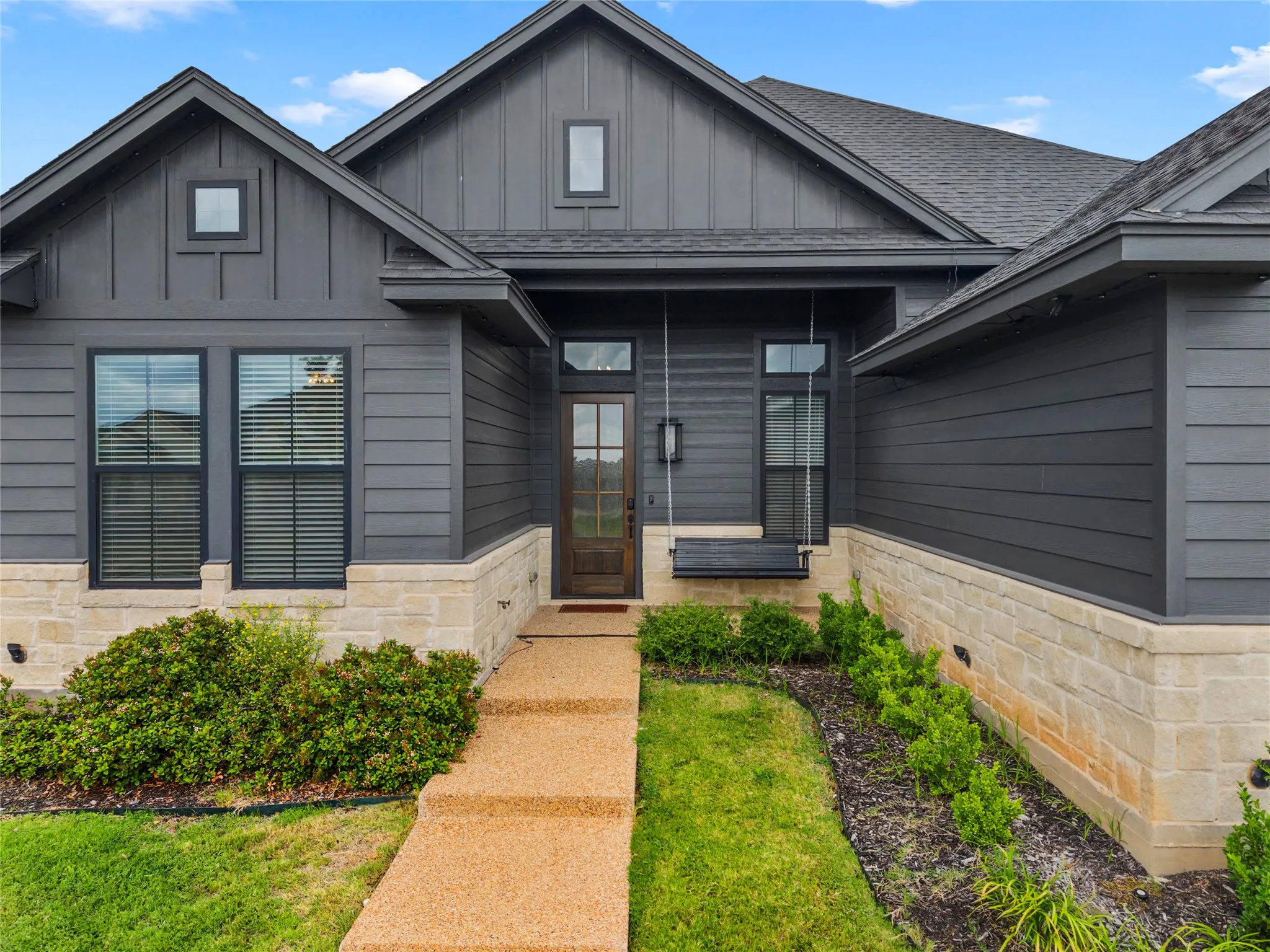 Architectural facade featuring dark horizontal siding and board-and-batten accents, stone veneer foundation, a gabled roofline, and an exposed aggregate walkway