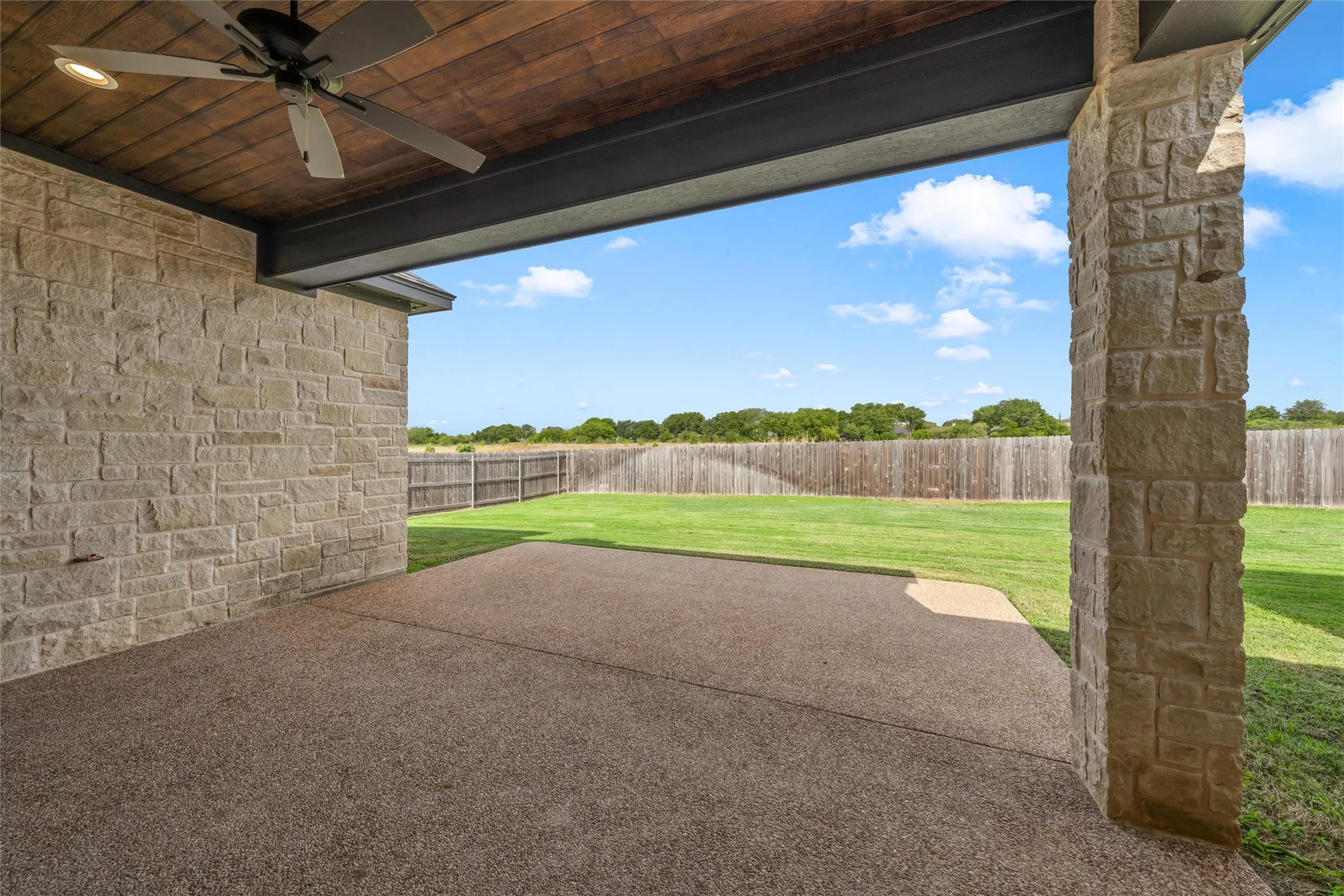 Covered patio with stone columns, exposed aggregate concrete flooring, wood-paneled ceiling, and a ceiling fan