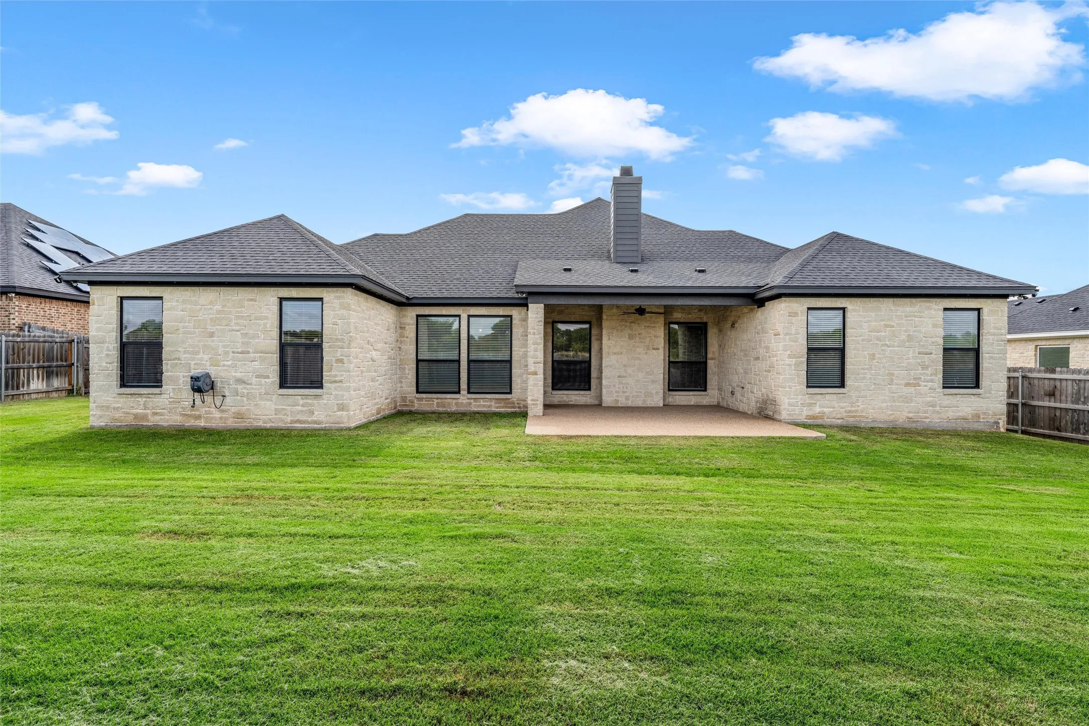 Extensive lawn space, stone exterior, black-framed windows, dark shingle roof, and a covered concrete patio