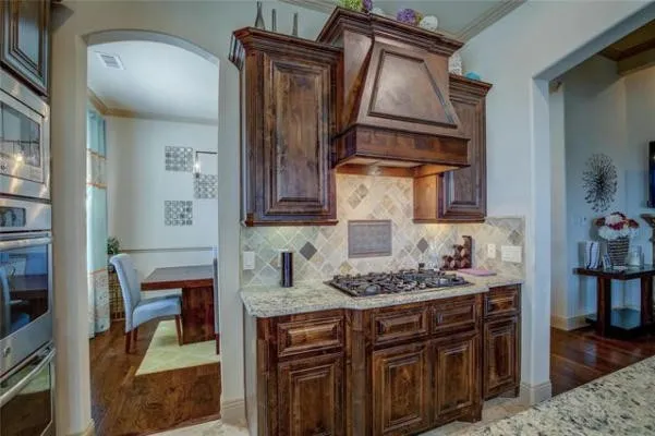 Dark wood cabinetry, built-in stainless steel double ovens, gas cooktop, granite countertops, and tile backsplash