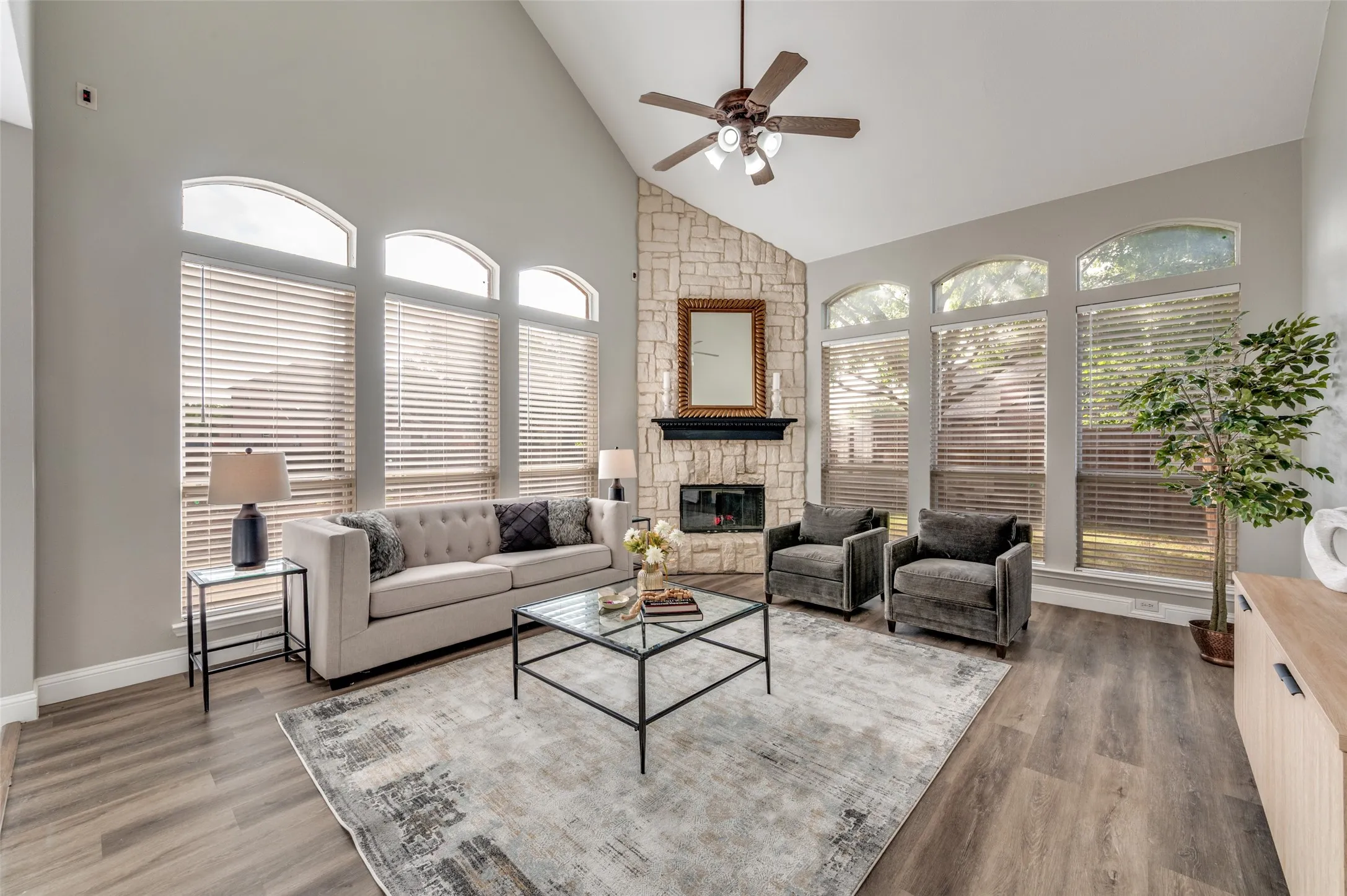 Vaulted ceiling, stone-clad fireplace, wood-finish flooring, multiple arched windows, and a ceiling fan with integrated lighting