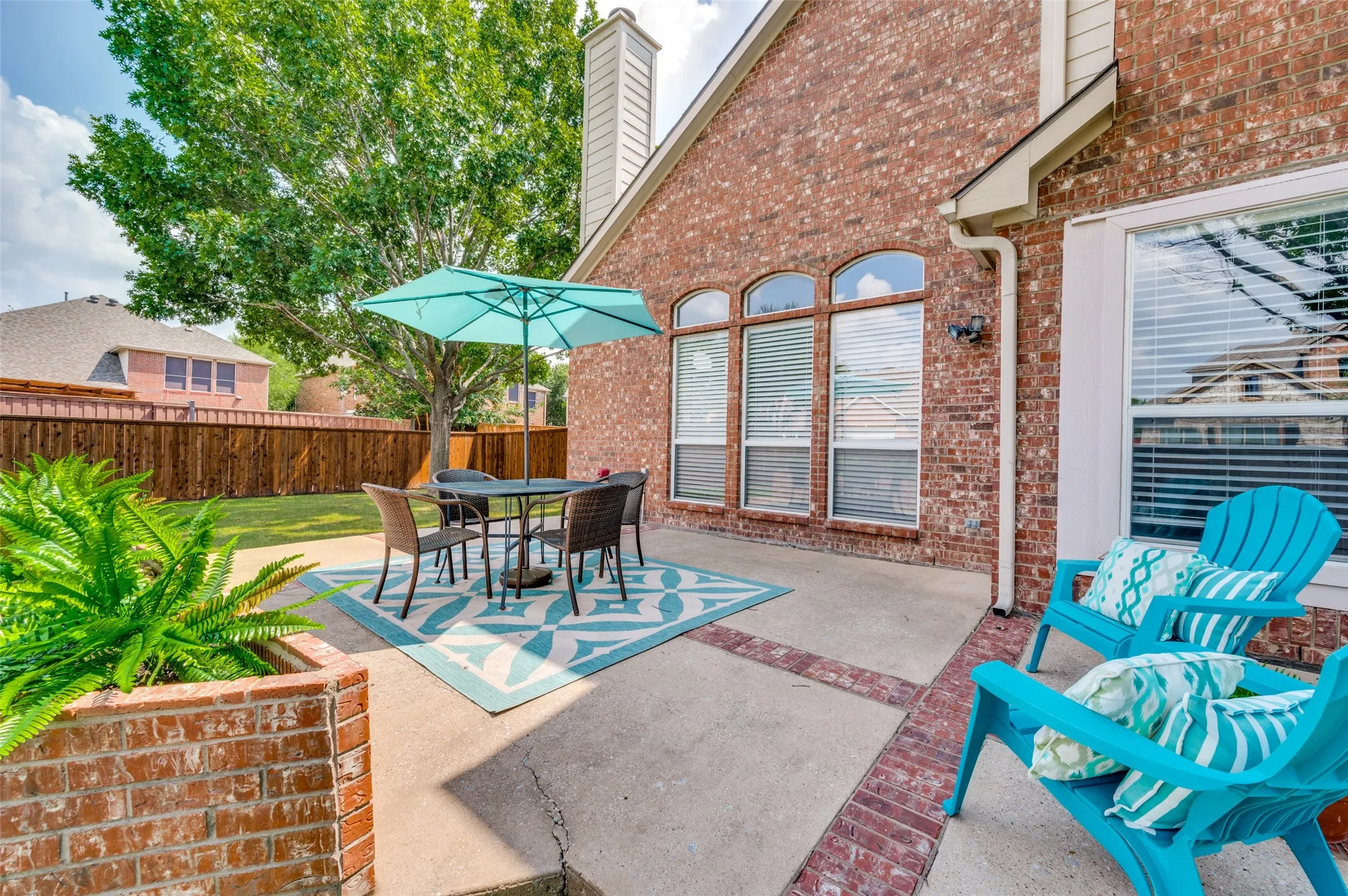 Expansive brick patio featuring mature landscaping, a built-in planter wall, and shaded seating area beneath a turquoise umbrella
