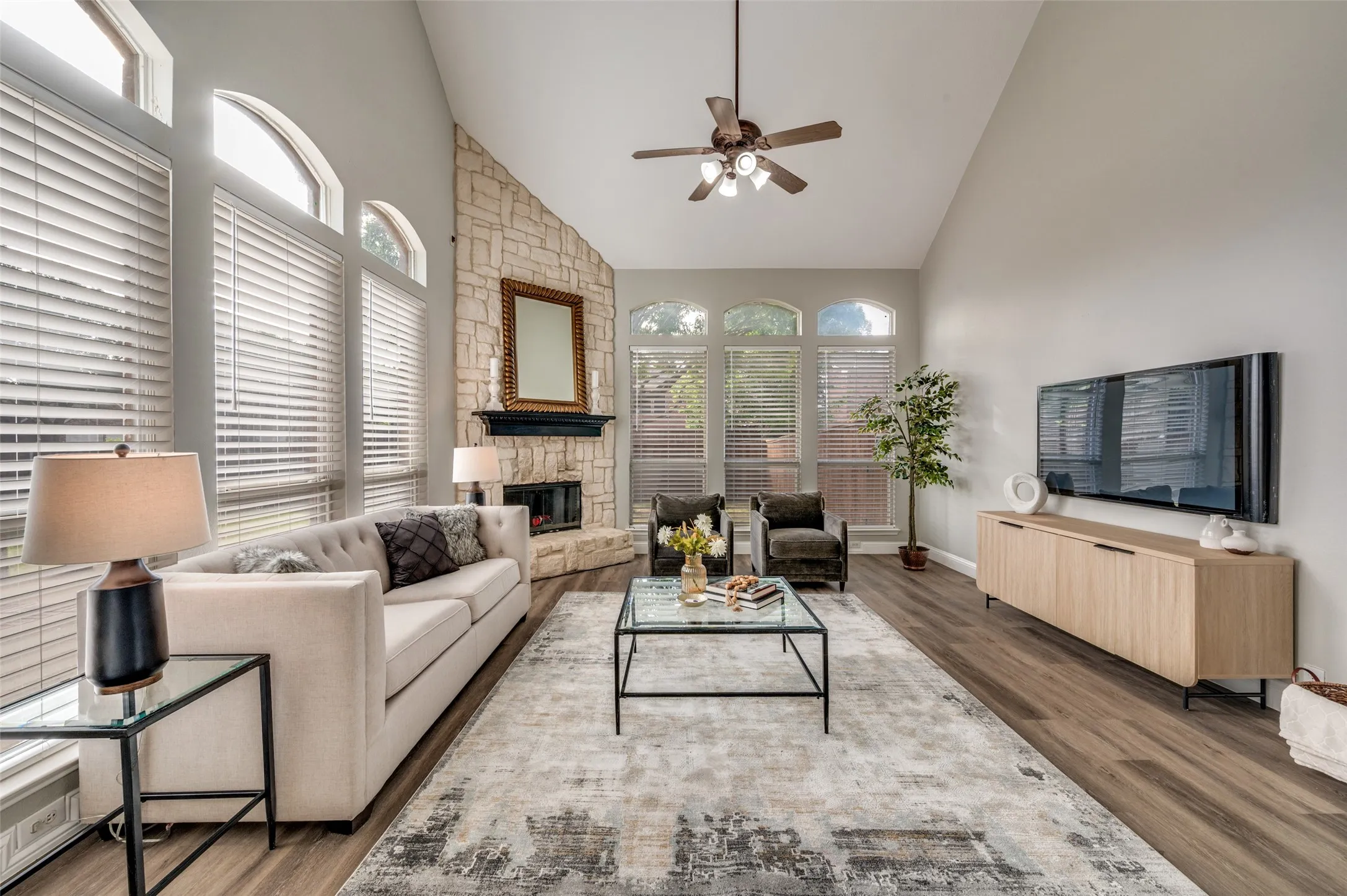 Vaulted ceiling with a ceiling fan, stone-clad fireplace with a mantel, multiple arched windows, and wood-finish flooring