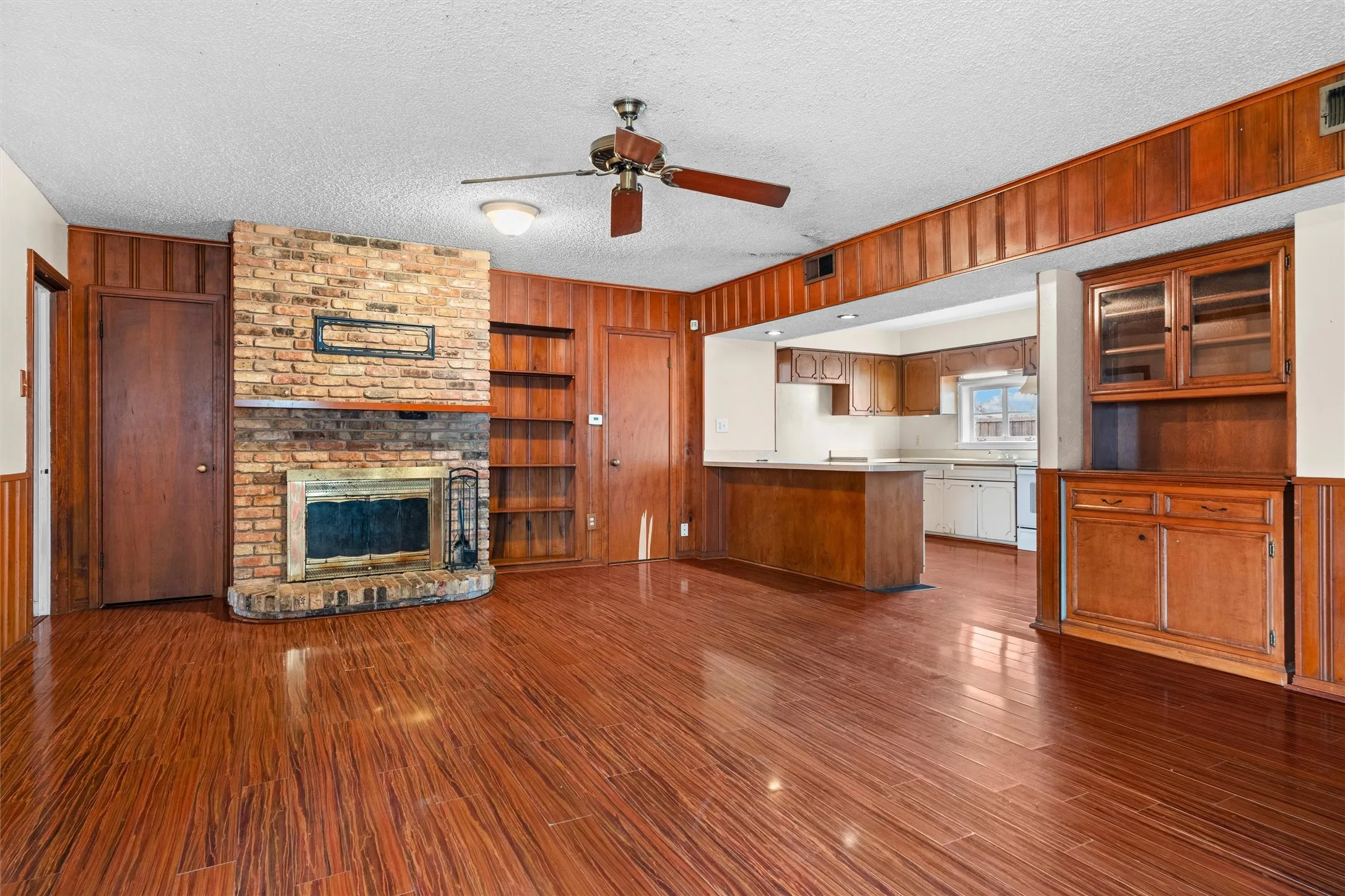 Brick-clad fireplace with hearth, wood-finish flooring, built-in shelving, wood wall paneling, and a ceiling fan
