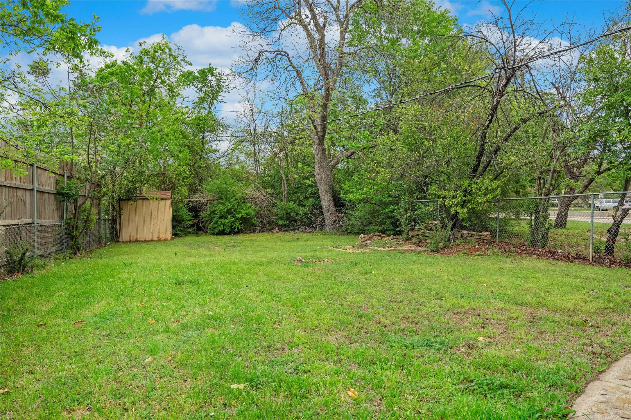 Expansive green lawn area with mature perimeter trees, a wood privacy fence, a chain-link fence, and a wood-sided shed