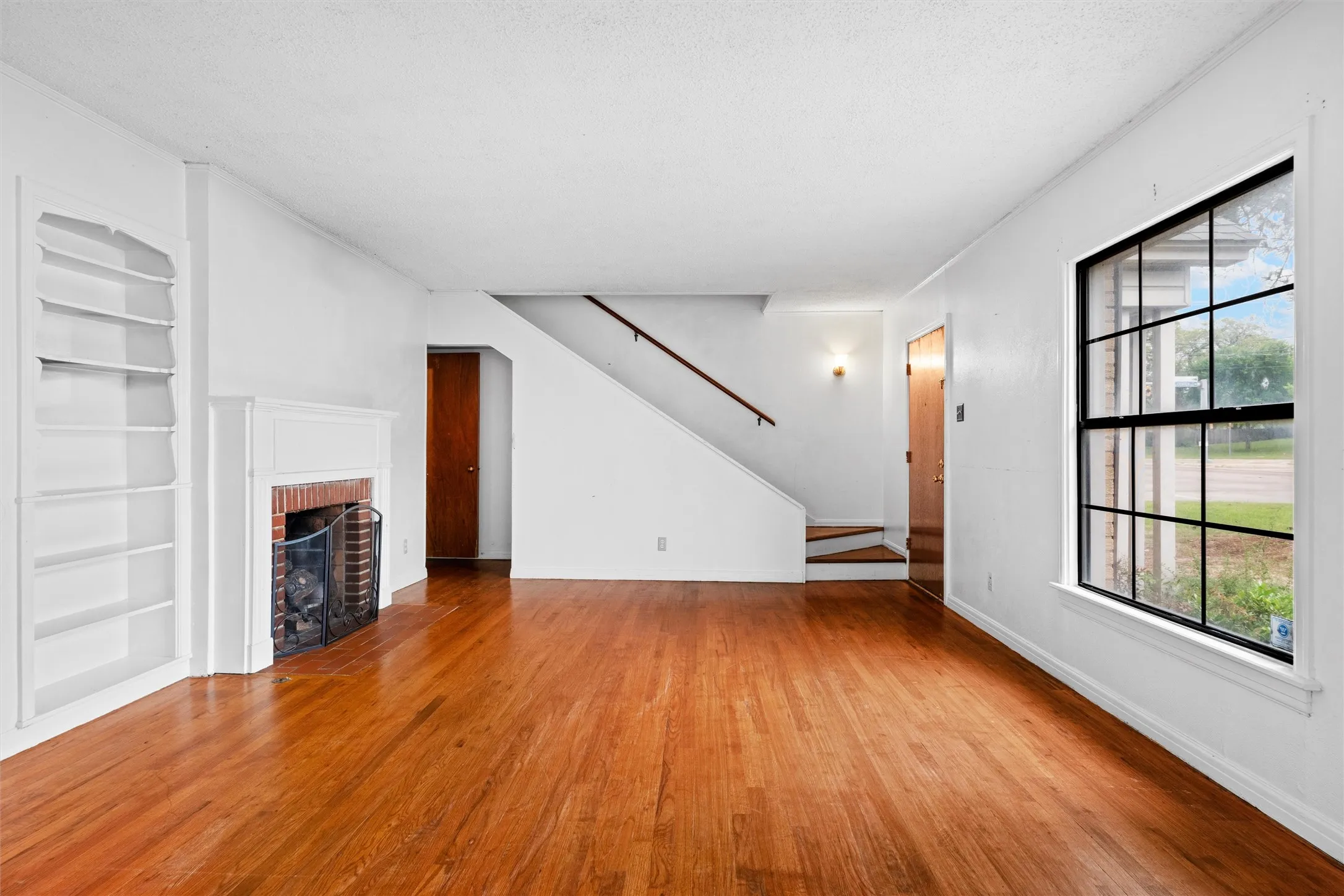 Hardwood flooring, brick fireplace with white mantel, built-in shelving, staircase with wood handrail, and a window with black grid panes