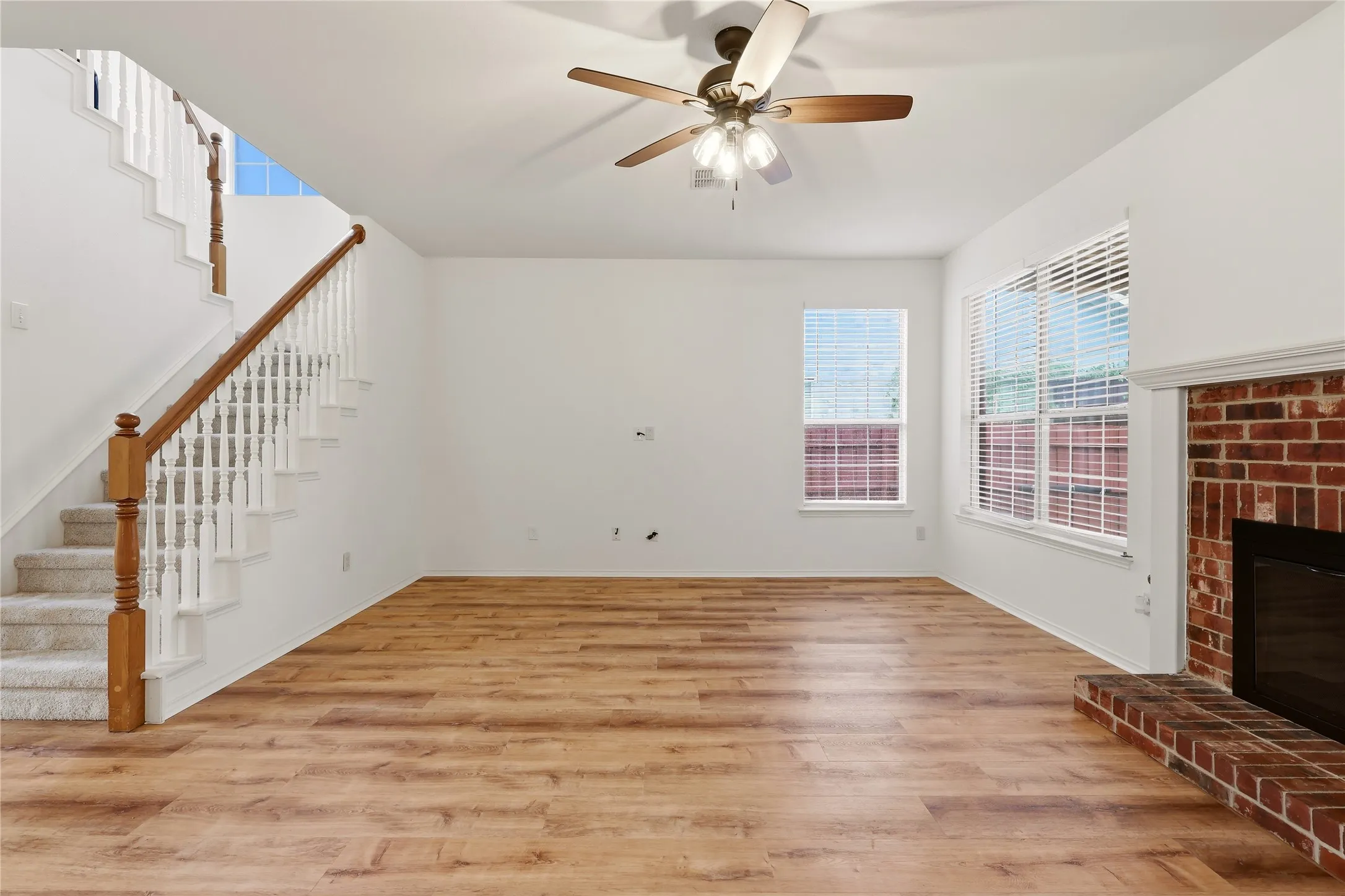 Hardwood-style flooring, brick-faced fireplace with hearth, ceiling fan with light fixture, and staircase with wood railing and carpeted treads