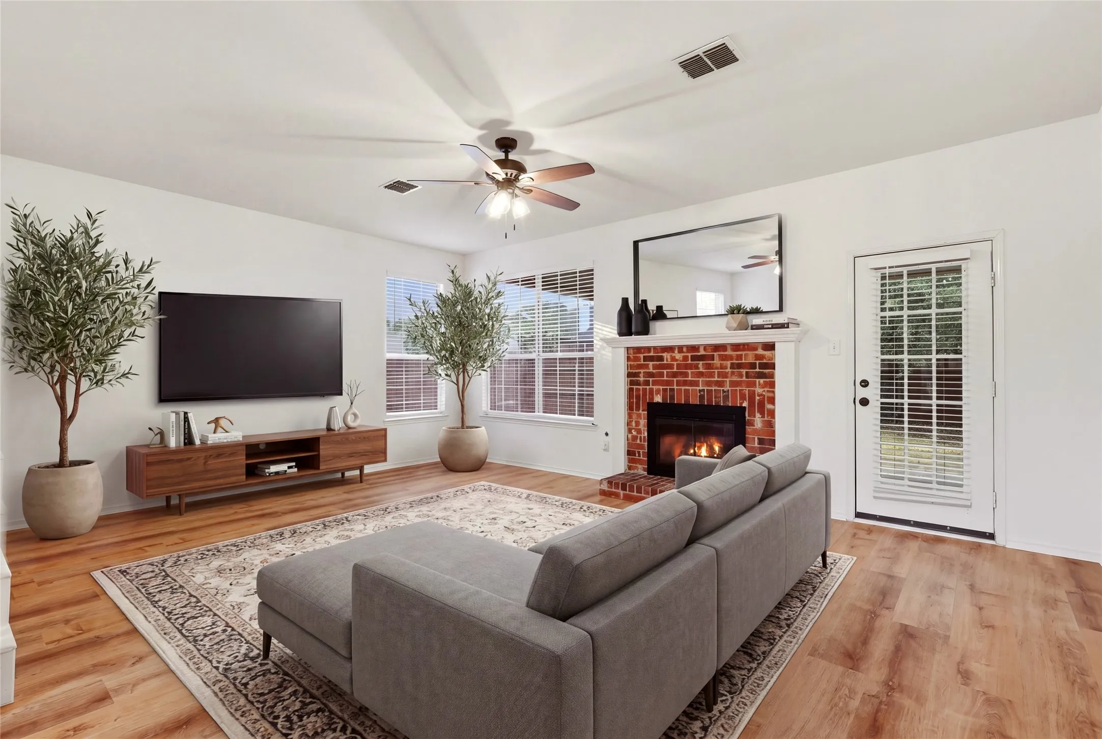 Open-concept living space with wood-finish flooring, a brick-surround fireplace with a white mantel, and ceiling fan.  Virtually staged.