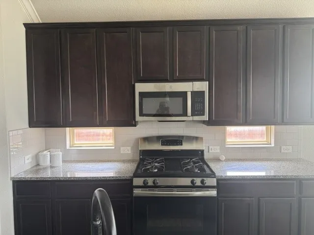 Dark wood-finish cabinetry, stainless steel microwave, stainless steel gas range, subway tile backsplash, and speckled countertops