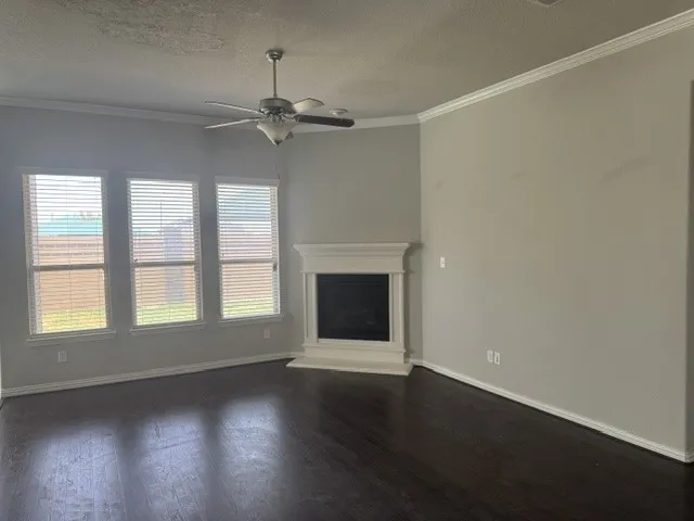 Wood-finish flooring, white crown molding, ceiling fan, three windows with blinds, and a white fireplace