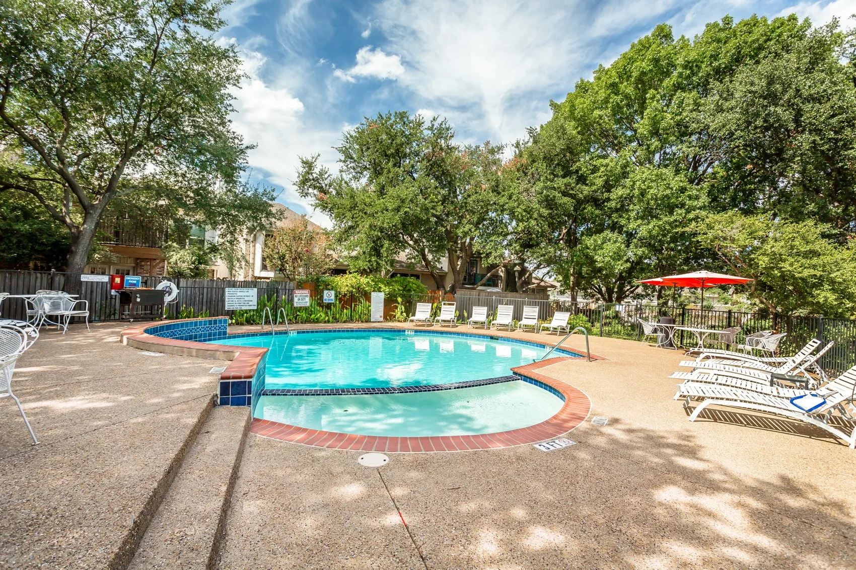 Curved swimming pool with blue and white tiling, surrounding hardscaping, and mature trees