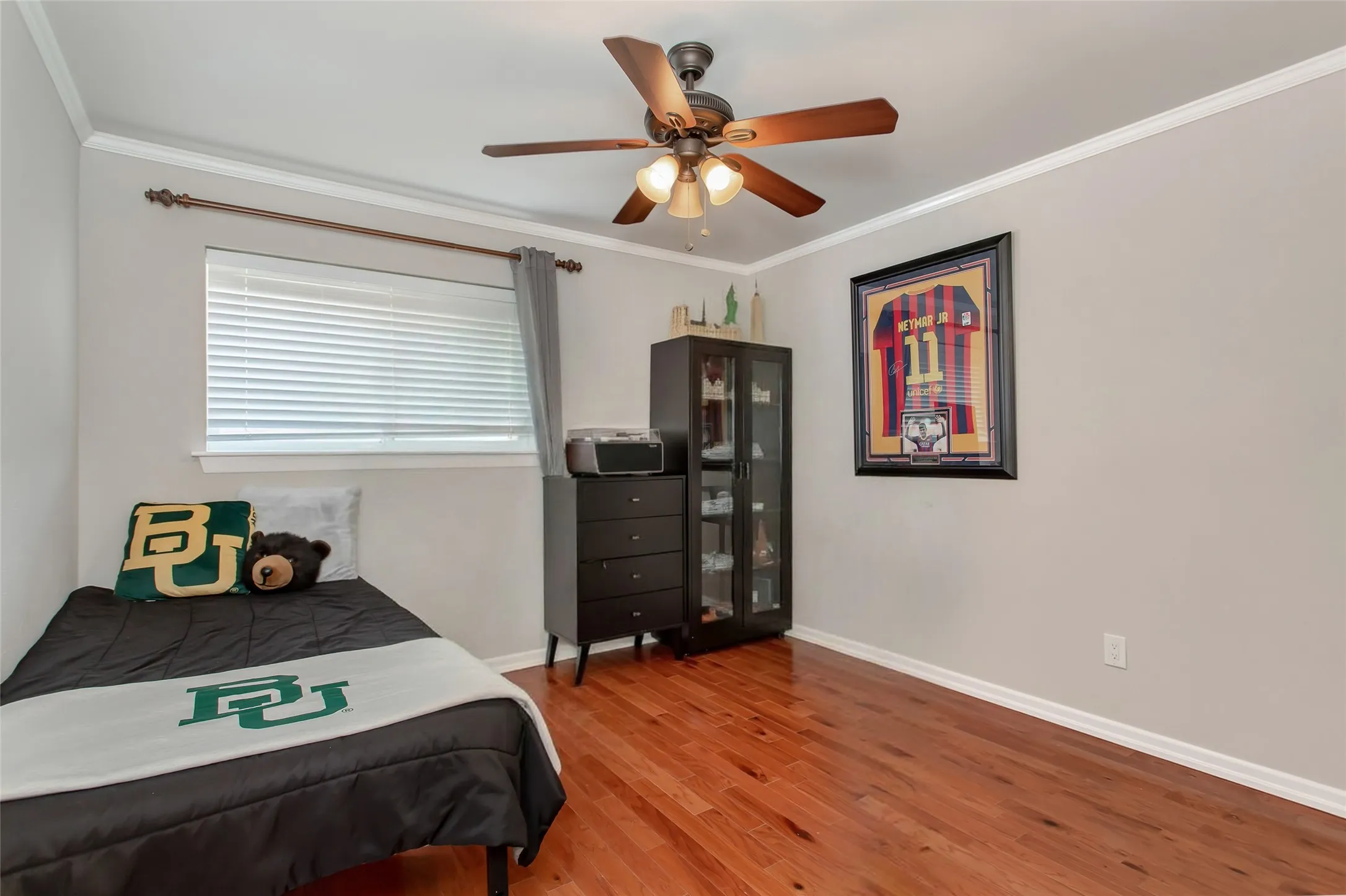 Bedroom featuring warm wood-finish flooring, ceiling fan with integrated lighting, large window with horizontal blinds, and dark wood cabinetry with glass-front storage
