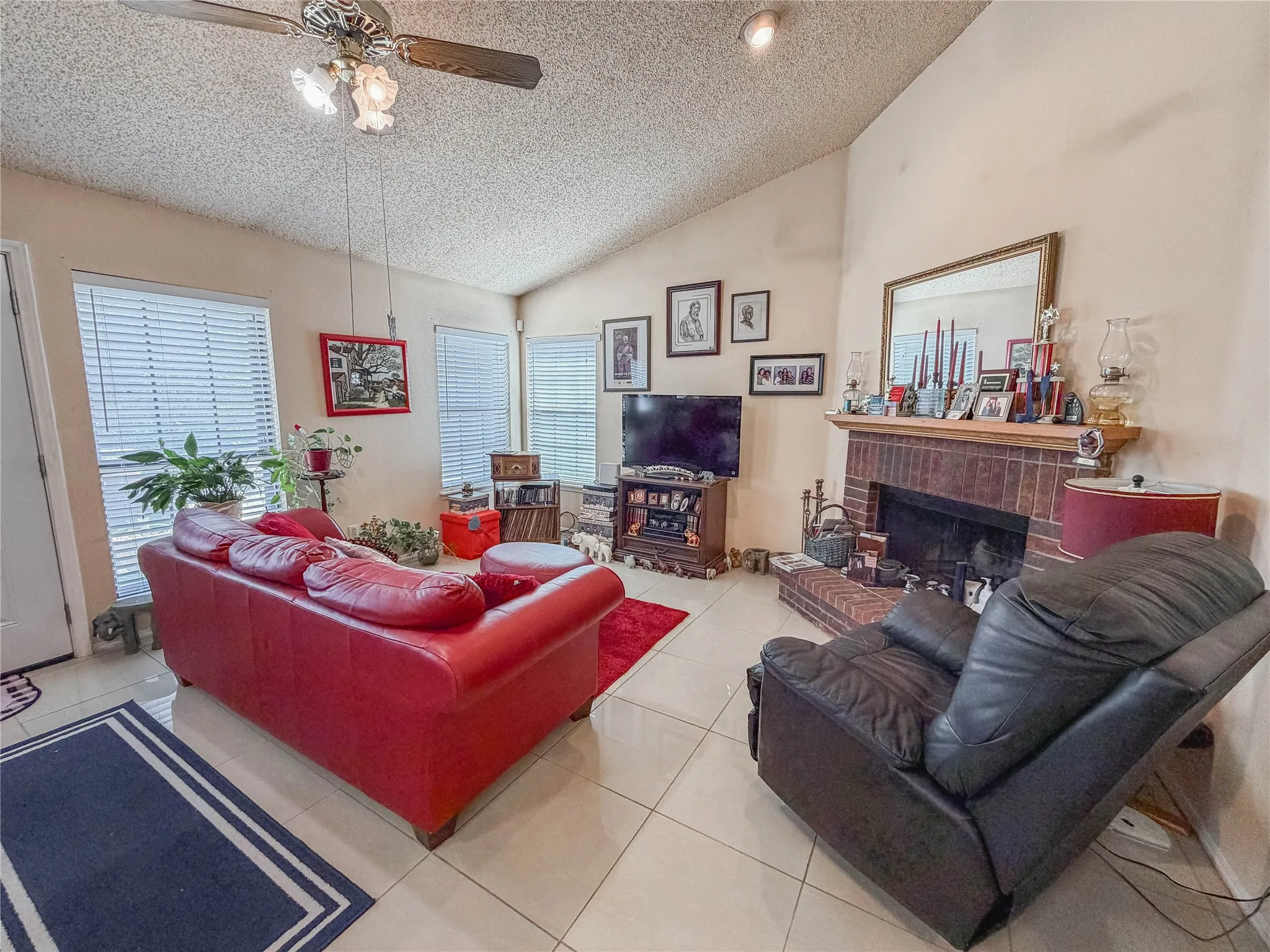 Vaulted ceiling, ceiling fan with integrated lighting, tiled flooring, brick-surround fireplace with wood mantel, multiple windows with blinds