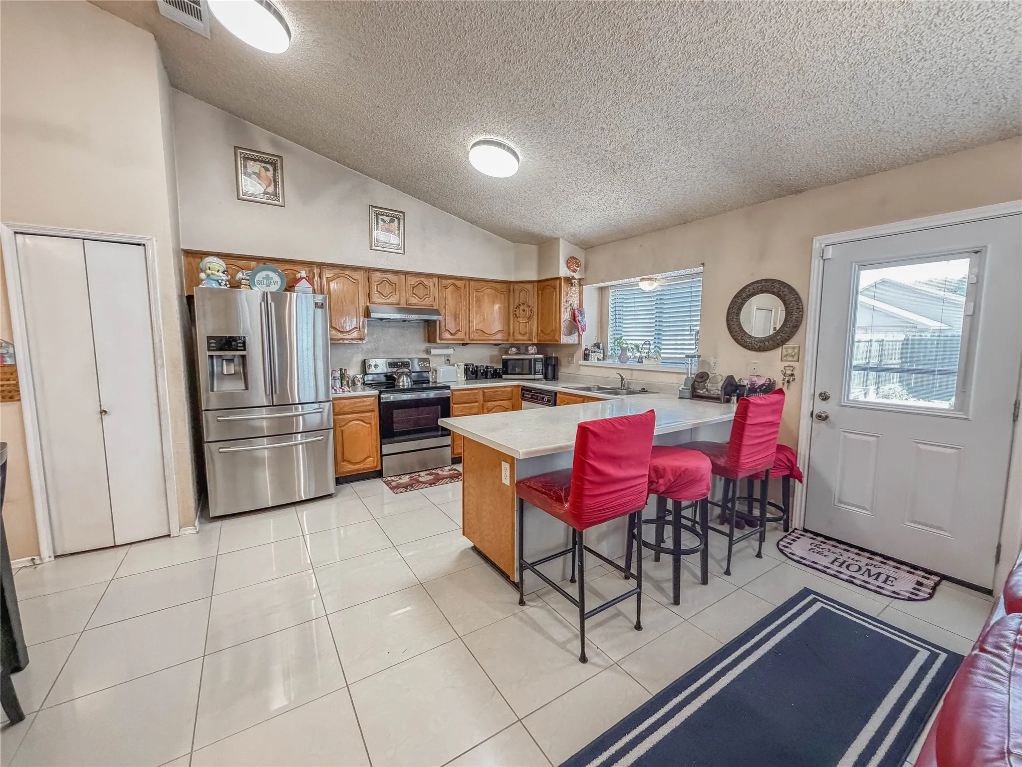 Kitchen featuring a vaulted ceiling, white tiled flooring, wood cabinetry, stainless steel appliances, and a breakfast bar with a light-colored countertop