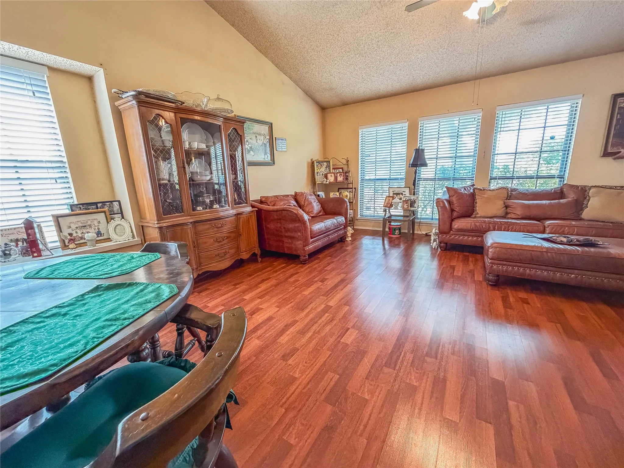 Vaulted ceiling, wood-finish flooring, multiple windows with blinds, and ceiling fan with lighting