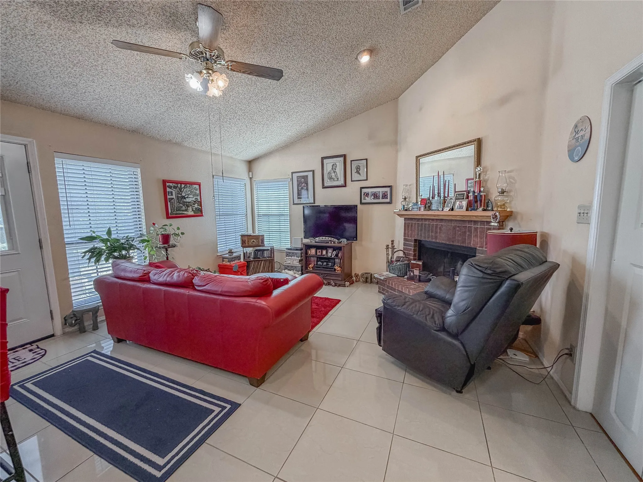 Vaulted ceiling with a ceiling fan, multiple windows with blinds, brick fireplace with a wood mantel, and large format tile flooring