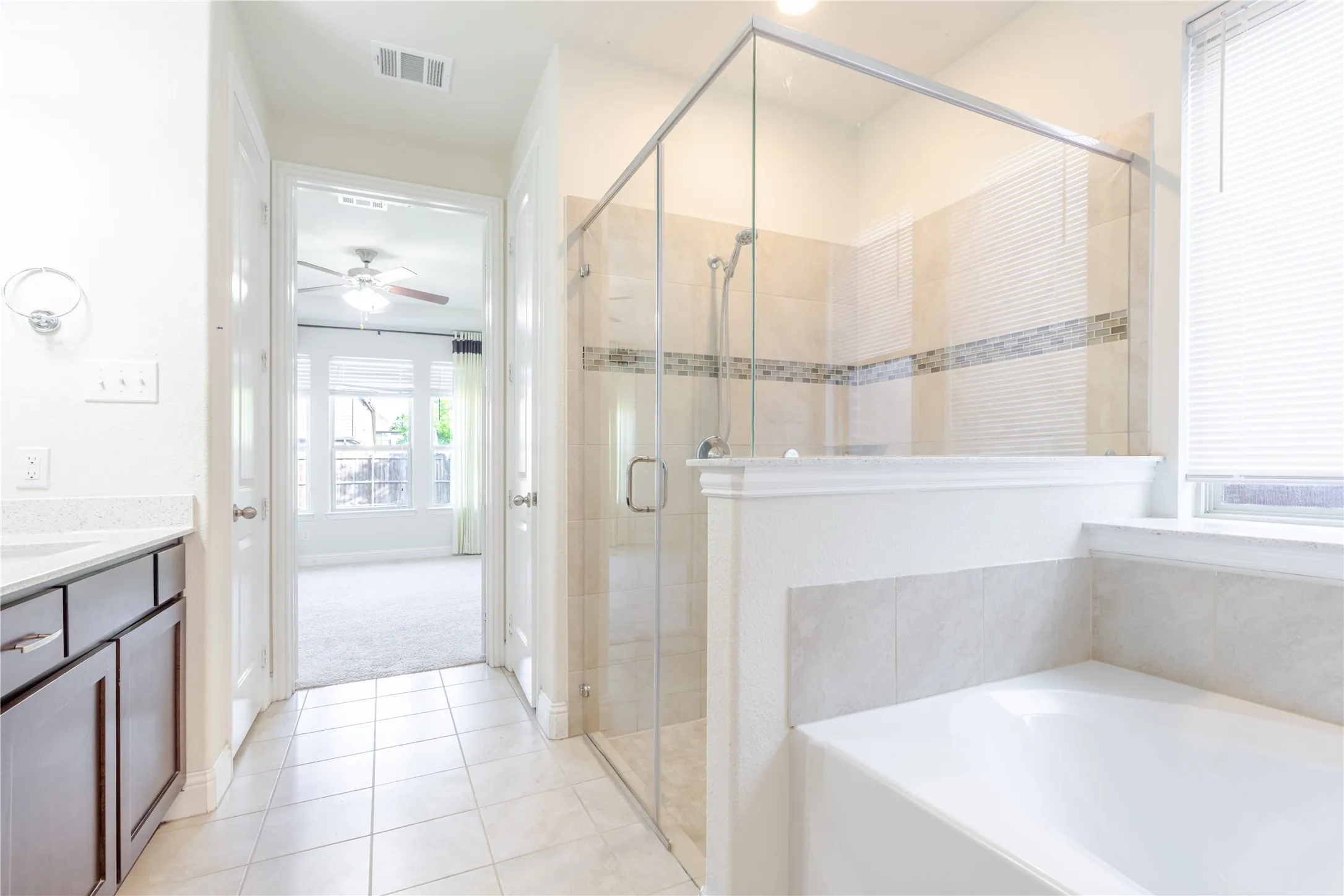 Spacious bathroom featuring a glass-enclosed shower with tile surround, a built-in tub, dark wood-finish vanity with a light-colored countertop, and light tile flooring