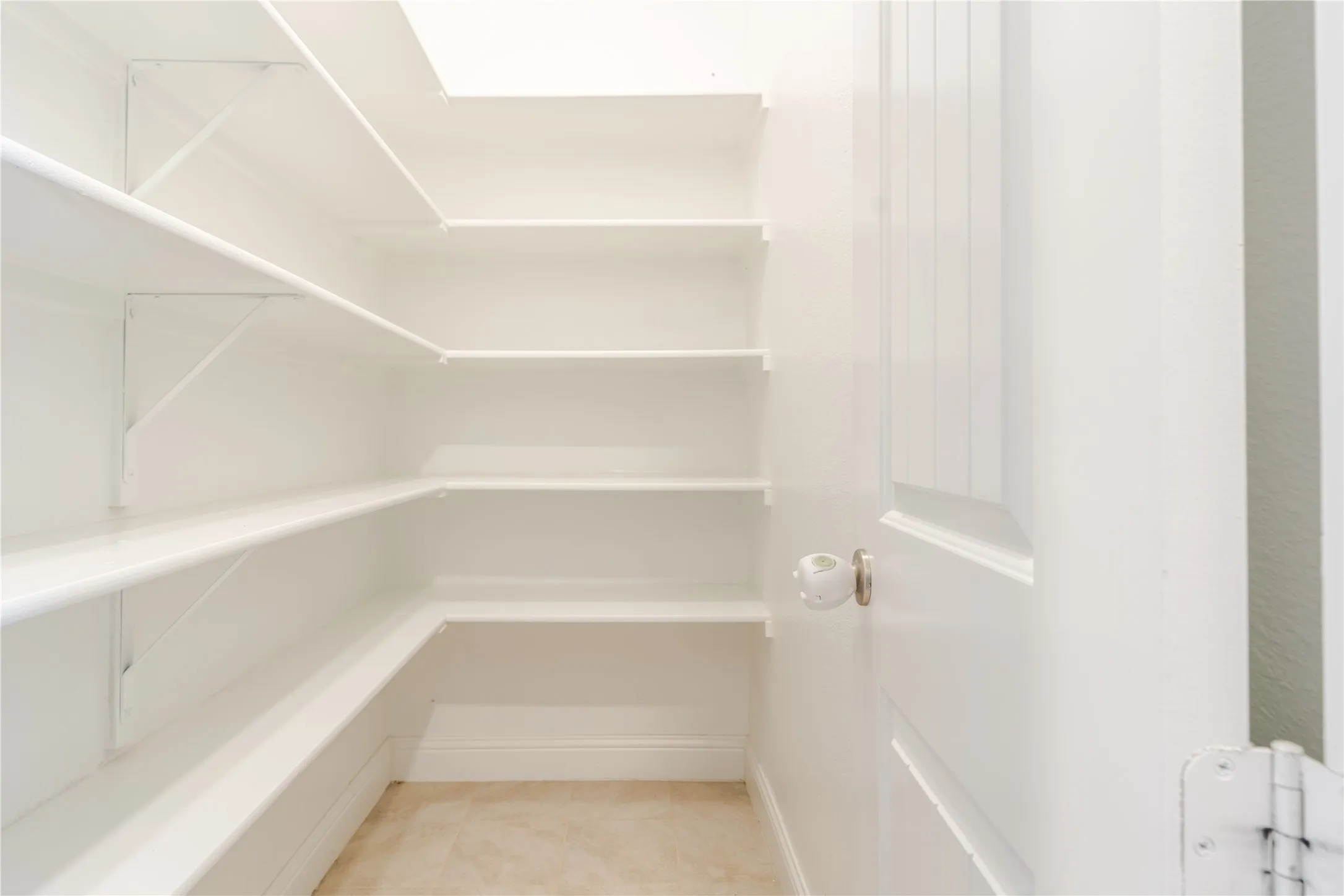 Walk-in pantry featuring white shelving, light neutral flooring, and a paneled door with a porcelain knob