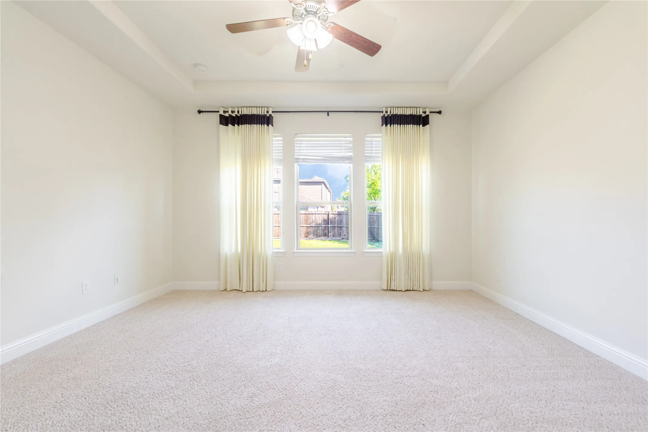 Spacious room featuring a tray ceiling, ceiling fan with light fixture, three windows with blinds and drapes, light-colored carpeting, and white baseboards