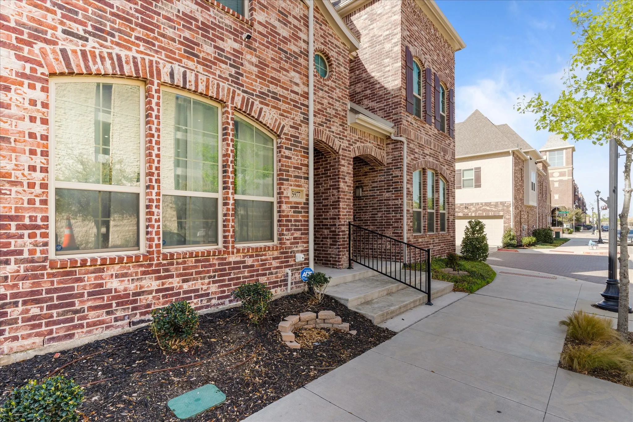 Brick facade featuring arched window headers, wrought iron railing, concrete steps, and landscaped beds
