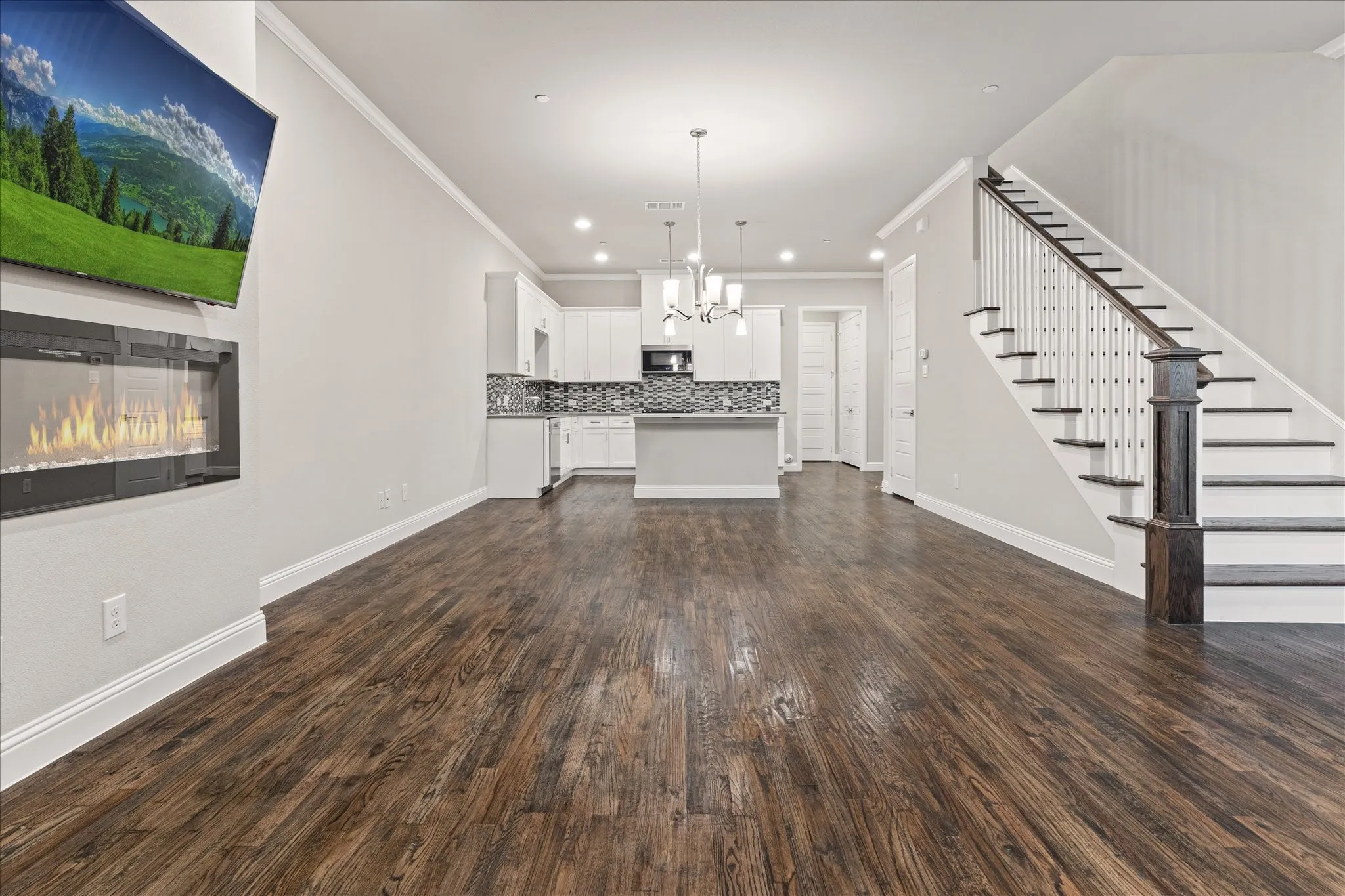 Spacious room featuring wood-finish flooring, an electric fireplace, a staircase with dark wood newel post and handrail, and crown molding
