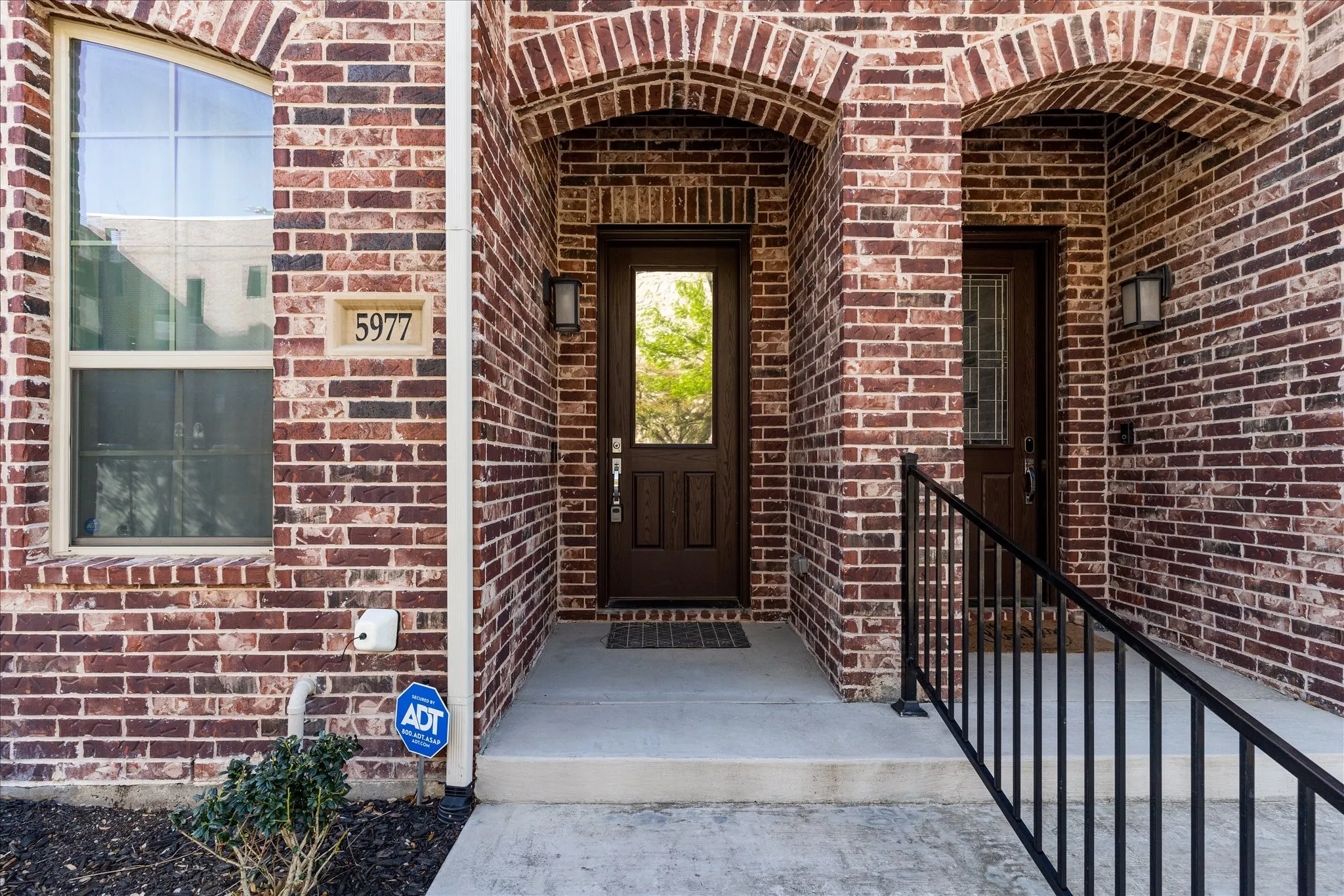 Brick facade with arched entryways, dark wood-finish entry door with glass panels, arched window, and concrete pathway with a black metal handrail