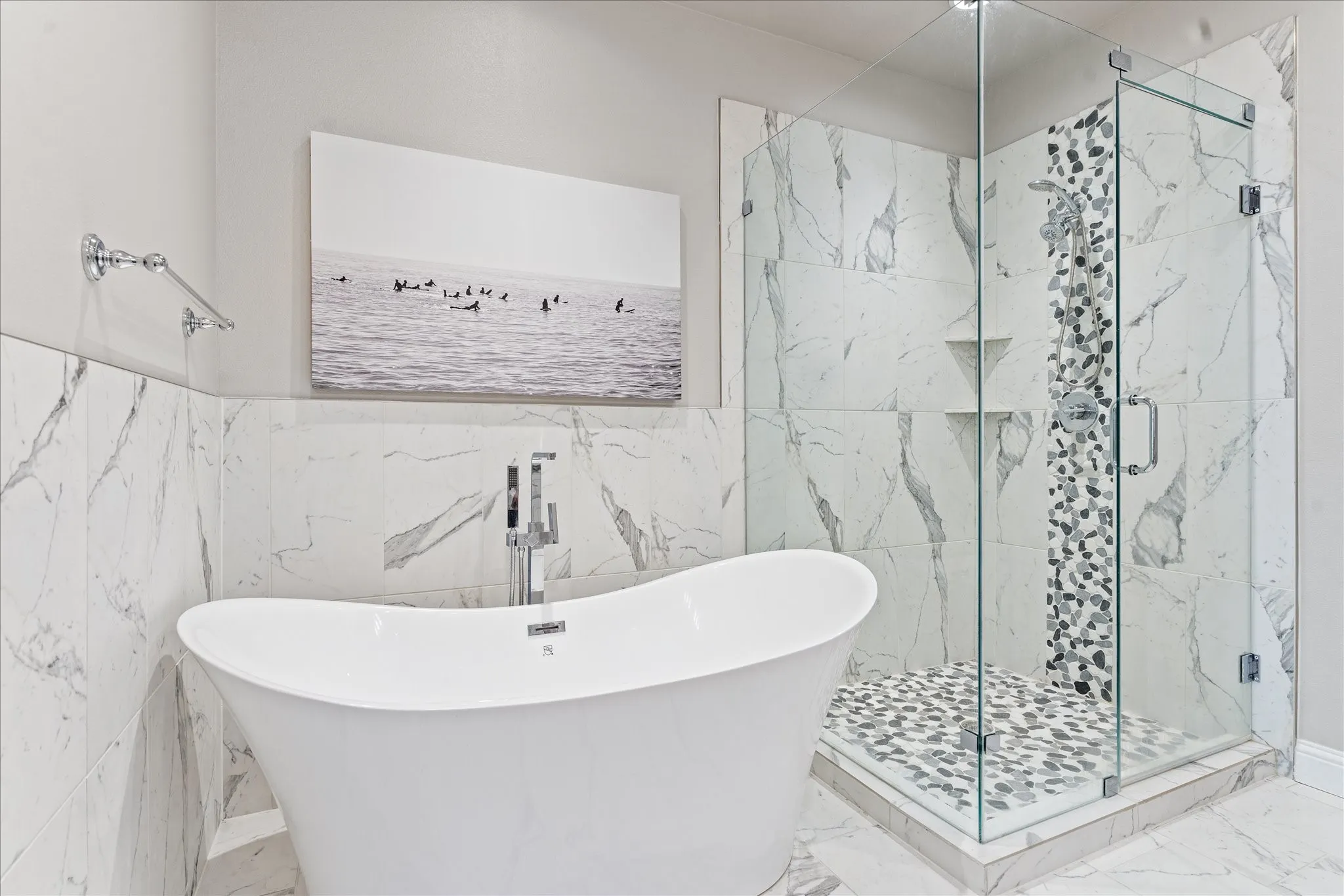 Freestanding soaking tub with chrome fixtures, glass-enclosed shower, stone-finish tile shower floor, stone-finish tile accent wall, and large format wall tile with a veined pattern