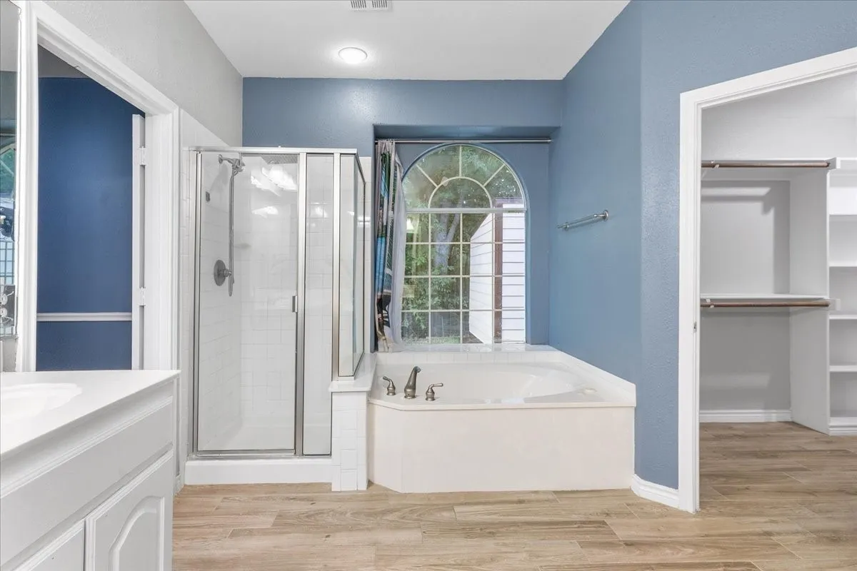 Bathroom featuring a corner soaking tub, an arched window, a glass-enclosed shower, a vanity with a white countertop, and wood-finish flooring