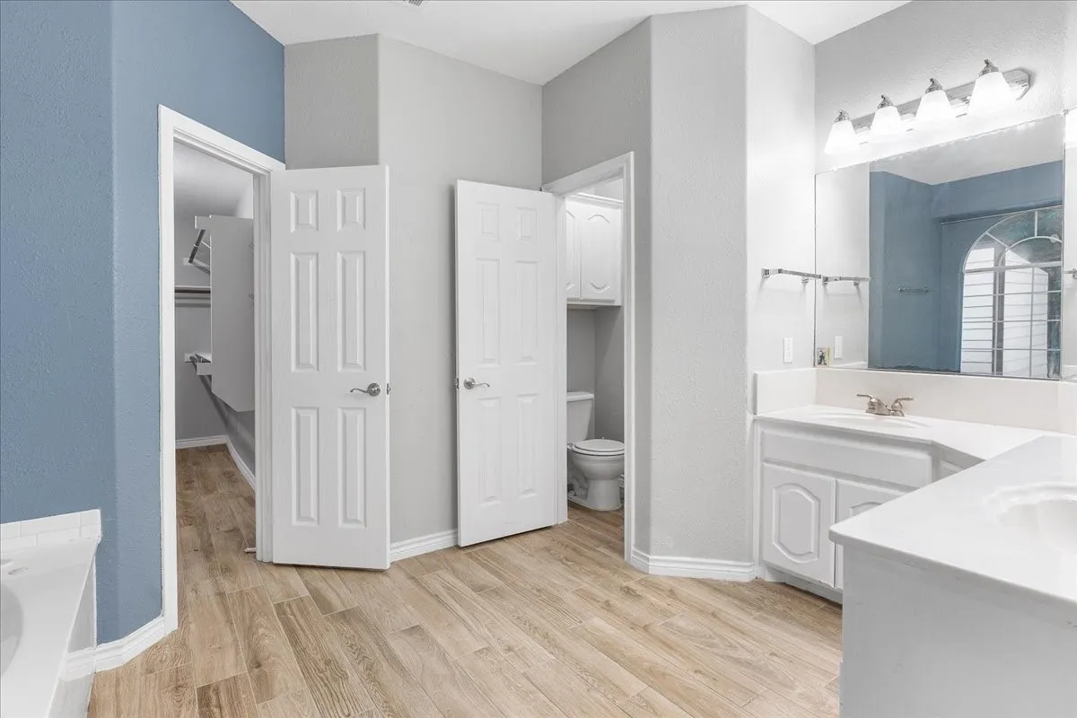 Bathroom featuring dual white vanities with integrated sinks, wood-finish flooring, and a mirrored wall with a five-light fixture