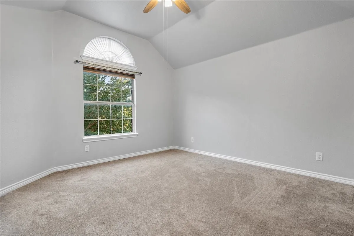 Spacious room featuring vaulted ceilings, neutral-toned carpeting, and a prominent arched window with a slatted blind