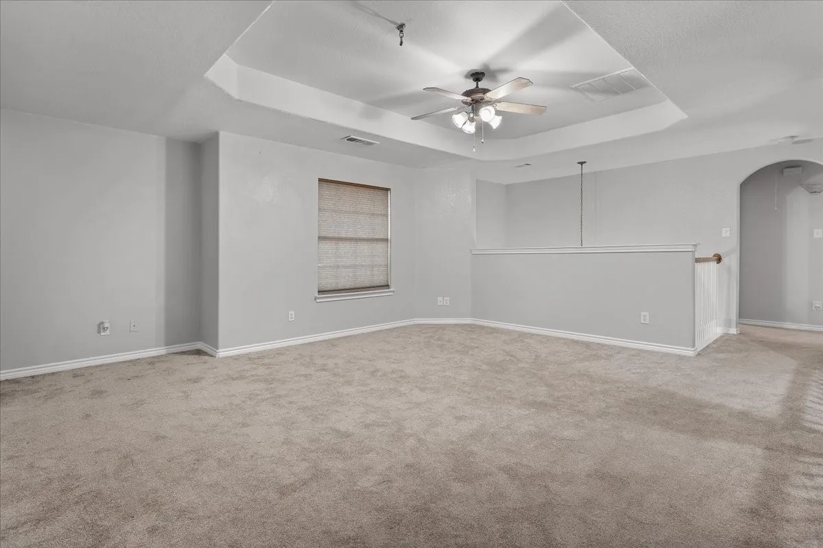 Spacious carpeted room featuring a tray ceiling with a ceiling fan, a window with blinds, an arched doorway, and a half-wall partition