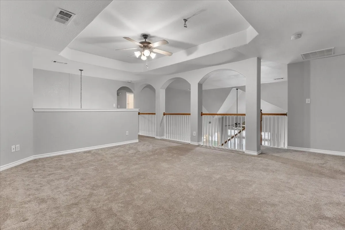 Spacious carpeted landing featuring a tray ceiling with a ceiling fan, white baluster railings with wood-finish handrails, and arched doorways