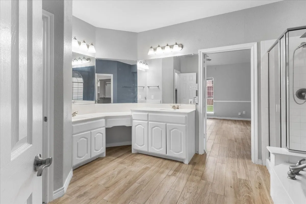 Bathroom featuring wood-finish flooring, dual vanity with white cabinetry, integrated sink basins, a glass-enclosed shower, and a built-in tub