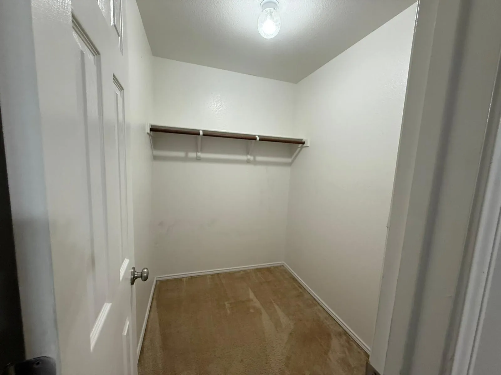Recessed closet with a single hanging rod and shelving, featuring a neutral carpeted floor and an overhead light fixture