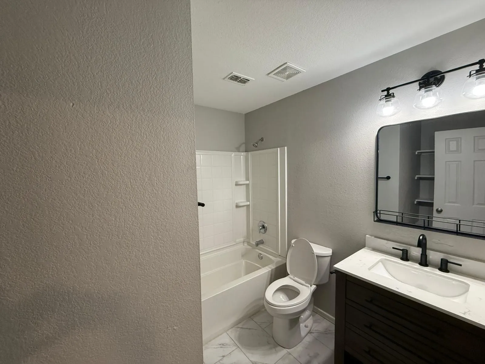 White bathtub with integrated shelving, a dark wood-finish vanity with a white countertop, an undermount sink, and a black-framed mirror
