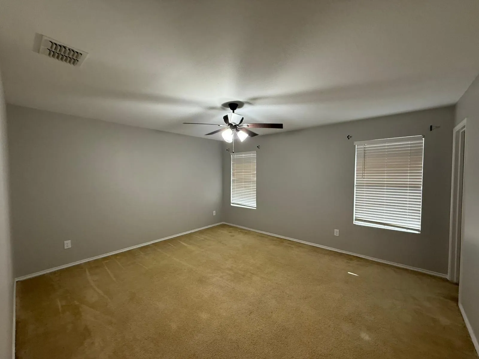 Spacious room with soft gold carpeting, neutral wall finishes, two windows with blinds, and a ceiling fan
