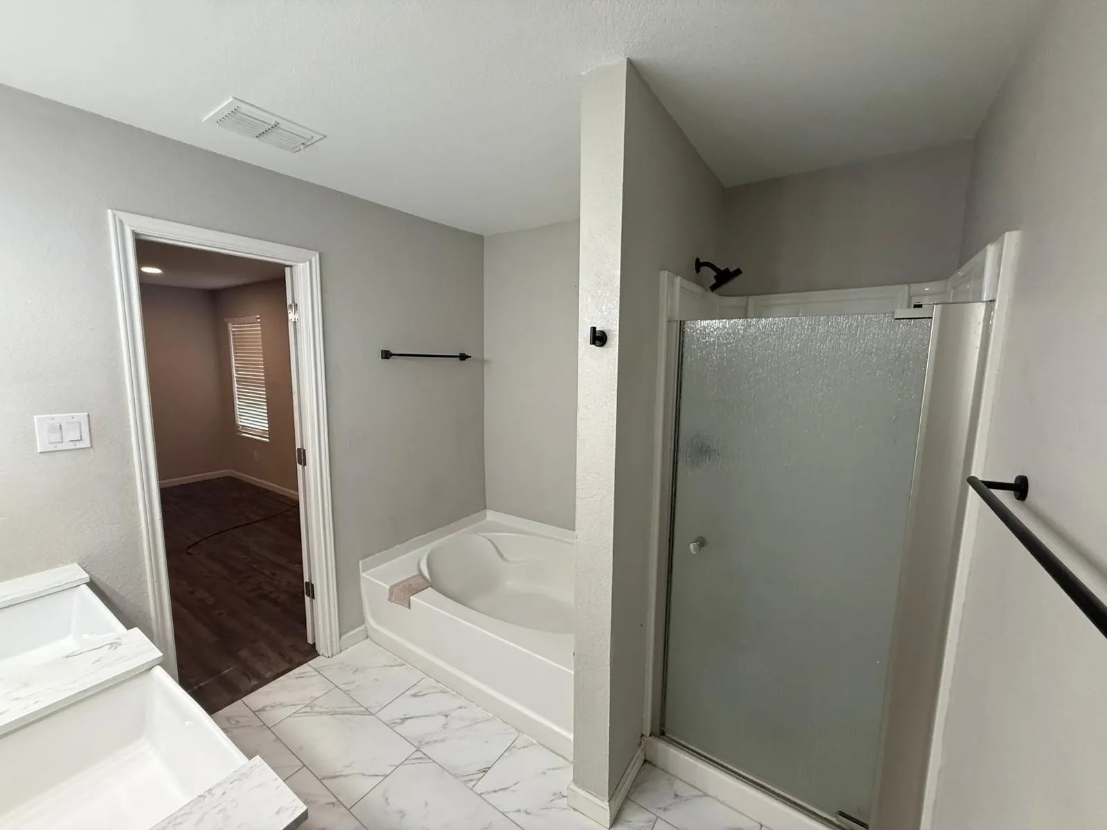White soaking tub, enclosed shower with frosted glass, marble-look tile flooring, and matte black hardware