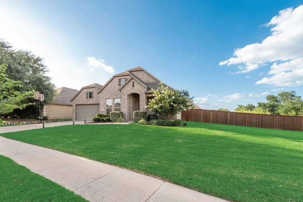 Brick exterior residence featuring a two-car garage, manicured lawn, established landscaping, a privacy fence, and a concrete sidewalk