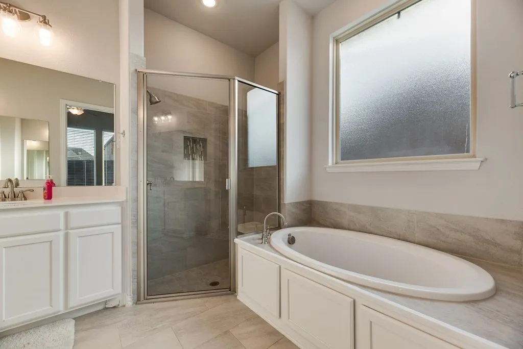 Oval soaking tub with tiled surround, frameless glass shower enclosure, single vanity with white cabinetry, and large frosted window