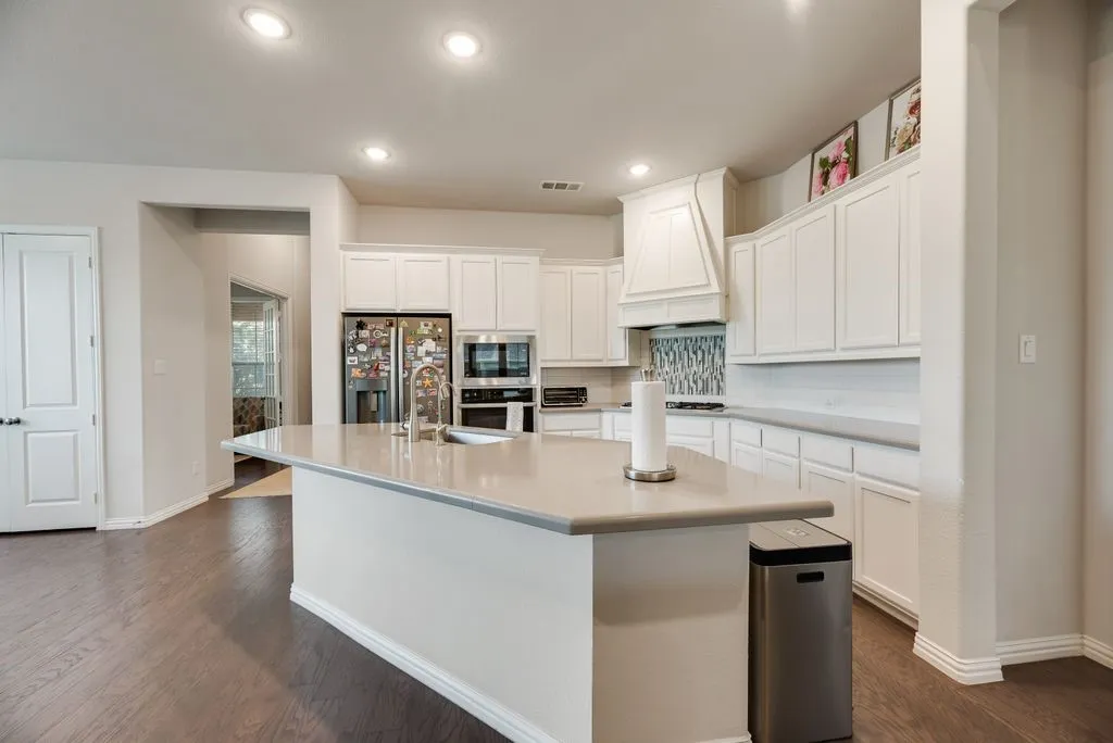 Kitchen featuring a substantial island with integrated sink, white cabinetry, integrated stainless steel appliances, a decorative range hood, and a tiled backsplash