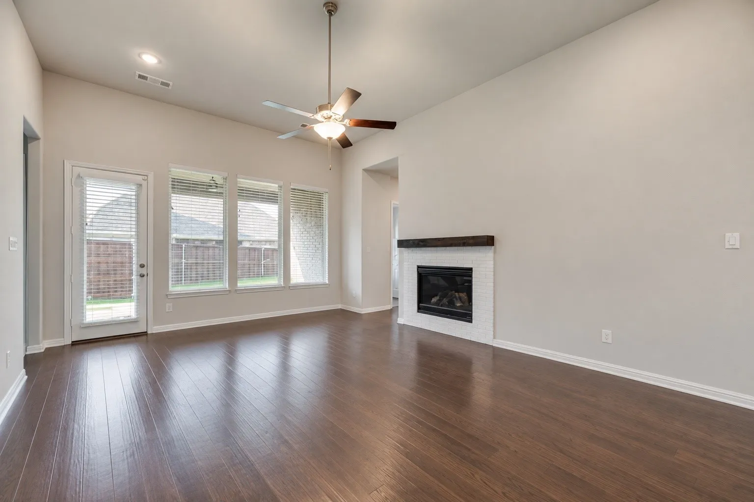 Vaulted ceilings with a ceiling fan, wood-finish flooring, a white brick fireplace with a wood mantel, and multiple windows with blinds