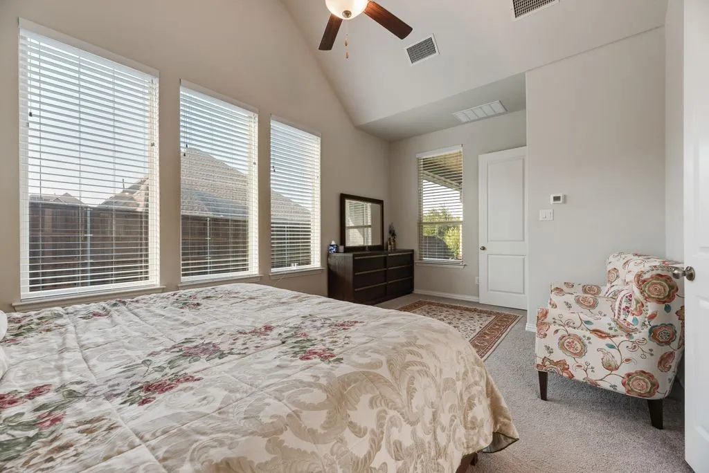 Vaulted ceiling bedroom featuring three large windows with blinds, a wood-finish ceiling fan, and light-colored carpeting