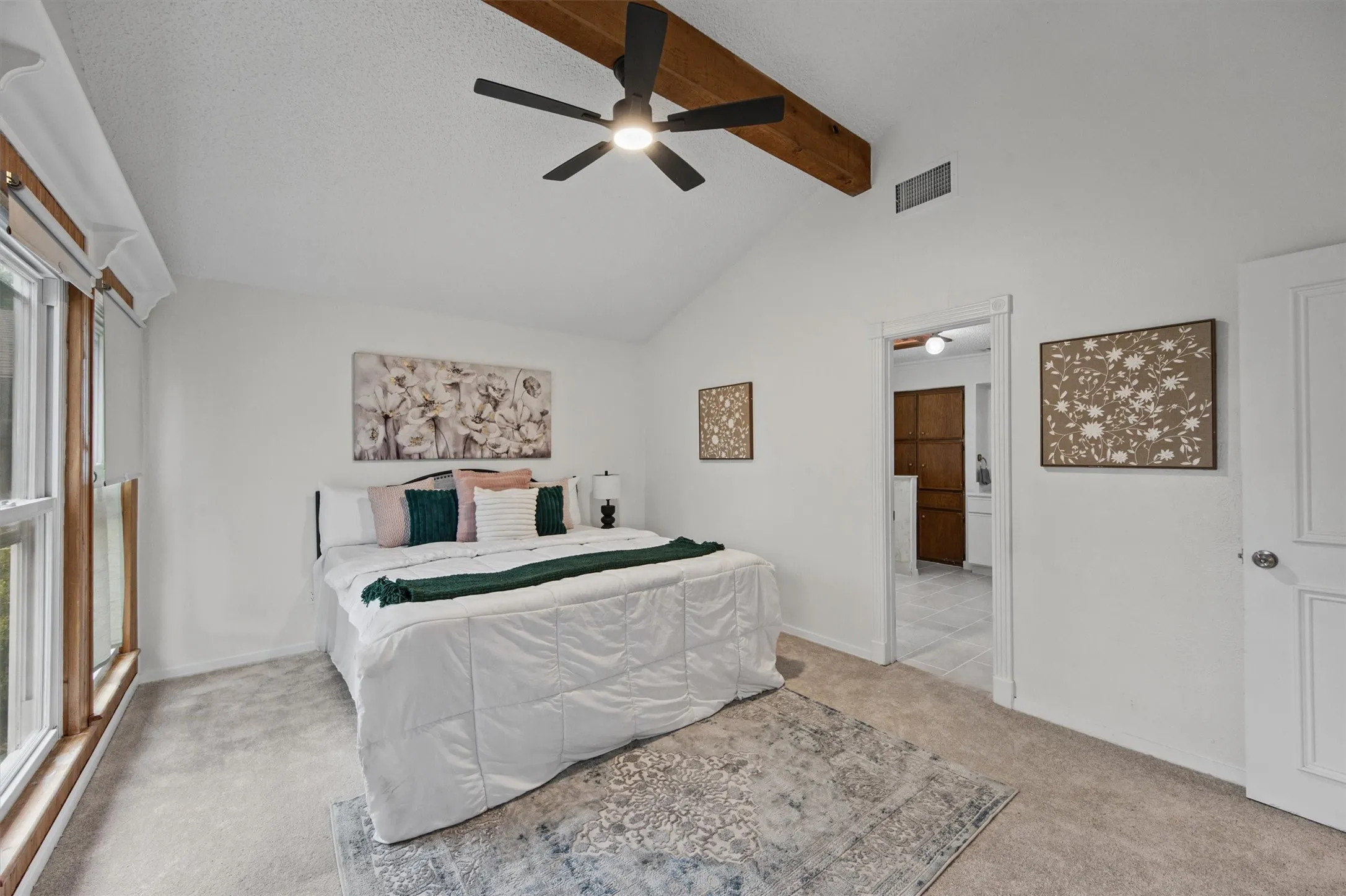 Vaulted ceiling with exposed wood beam, contemporary ceiling fan, light neutral carpeting, and a large window with wood trim