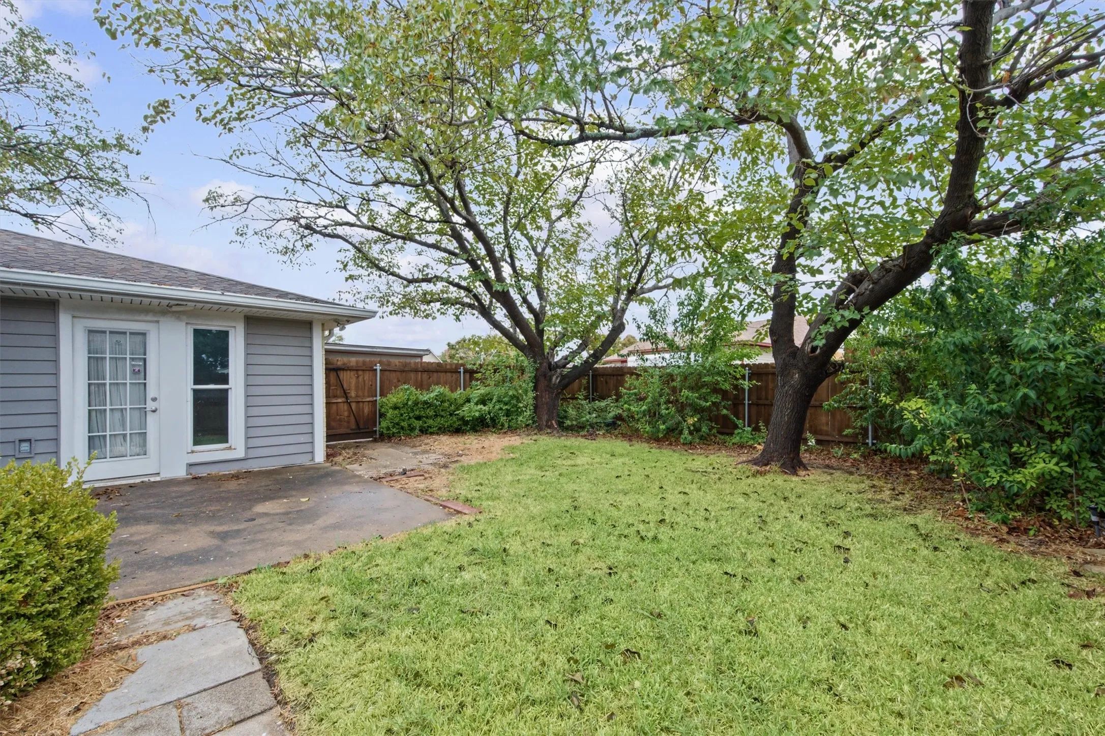 Backyard featuring a concrete patio, exterior siding, white French doors, mature trees, and a wood fence